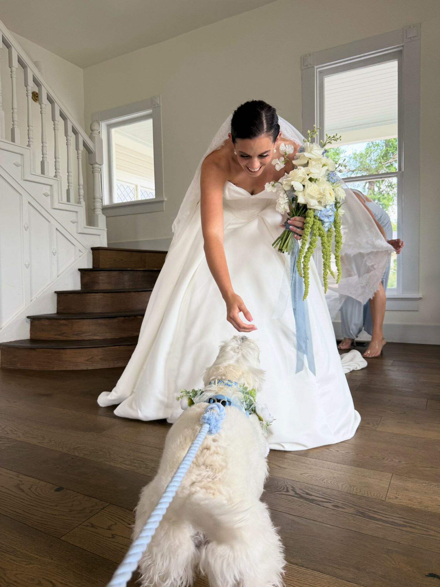Bride in wedding dress interacts with a dog inside a house, holding a bouquet of flowers, with stairs and windows in background.