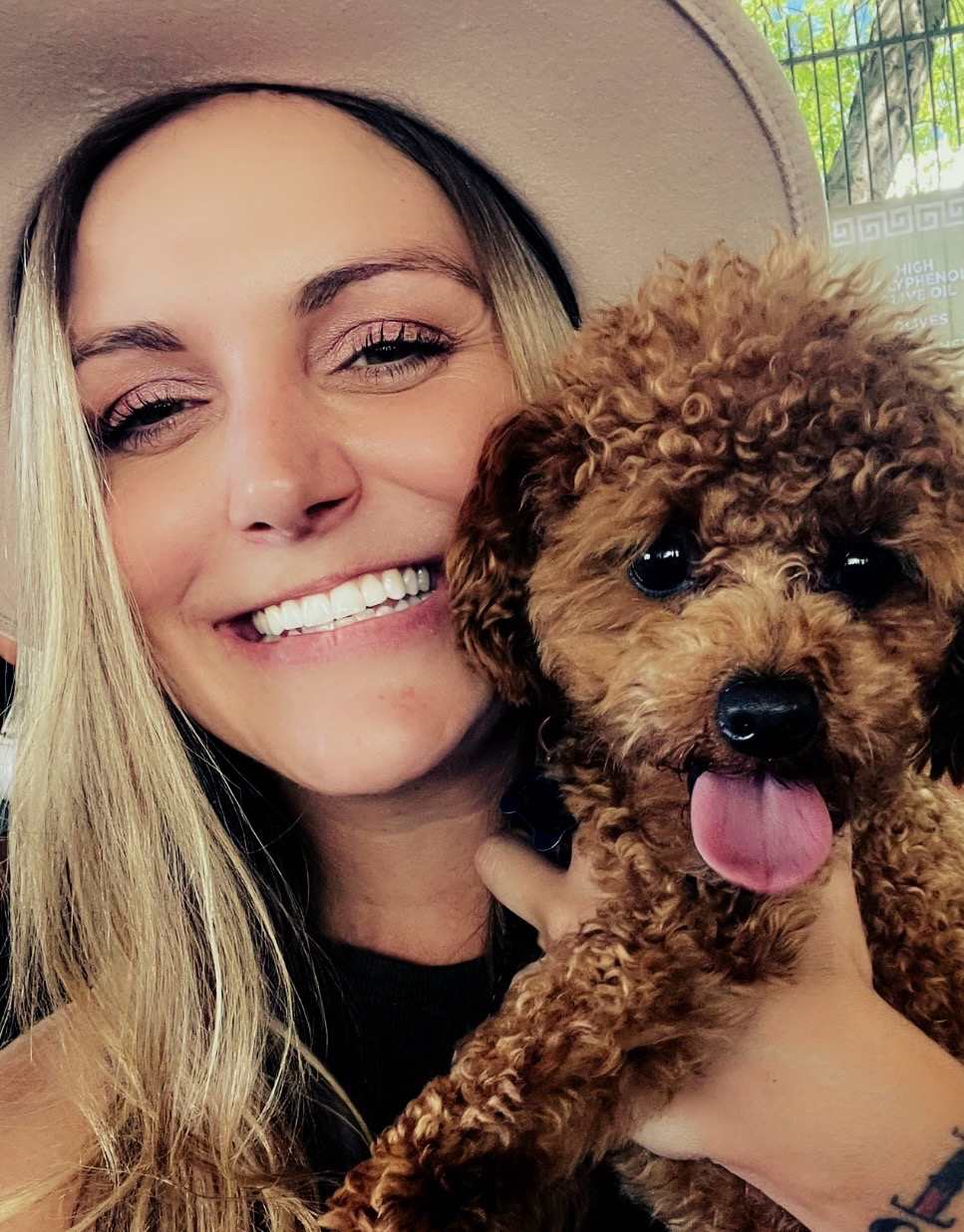 Woman with blonde hair smiling holding a small curly-haired dog with tongue out outdoors.