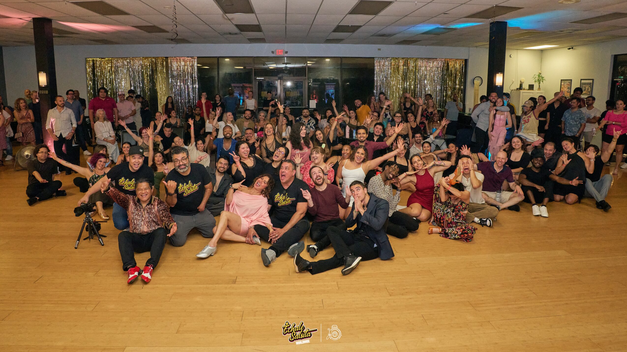Large group of people in a dance studio, sitting and standing, many raising hands, with a wooden floor and curtains in the background.