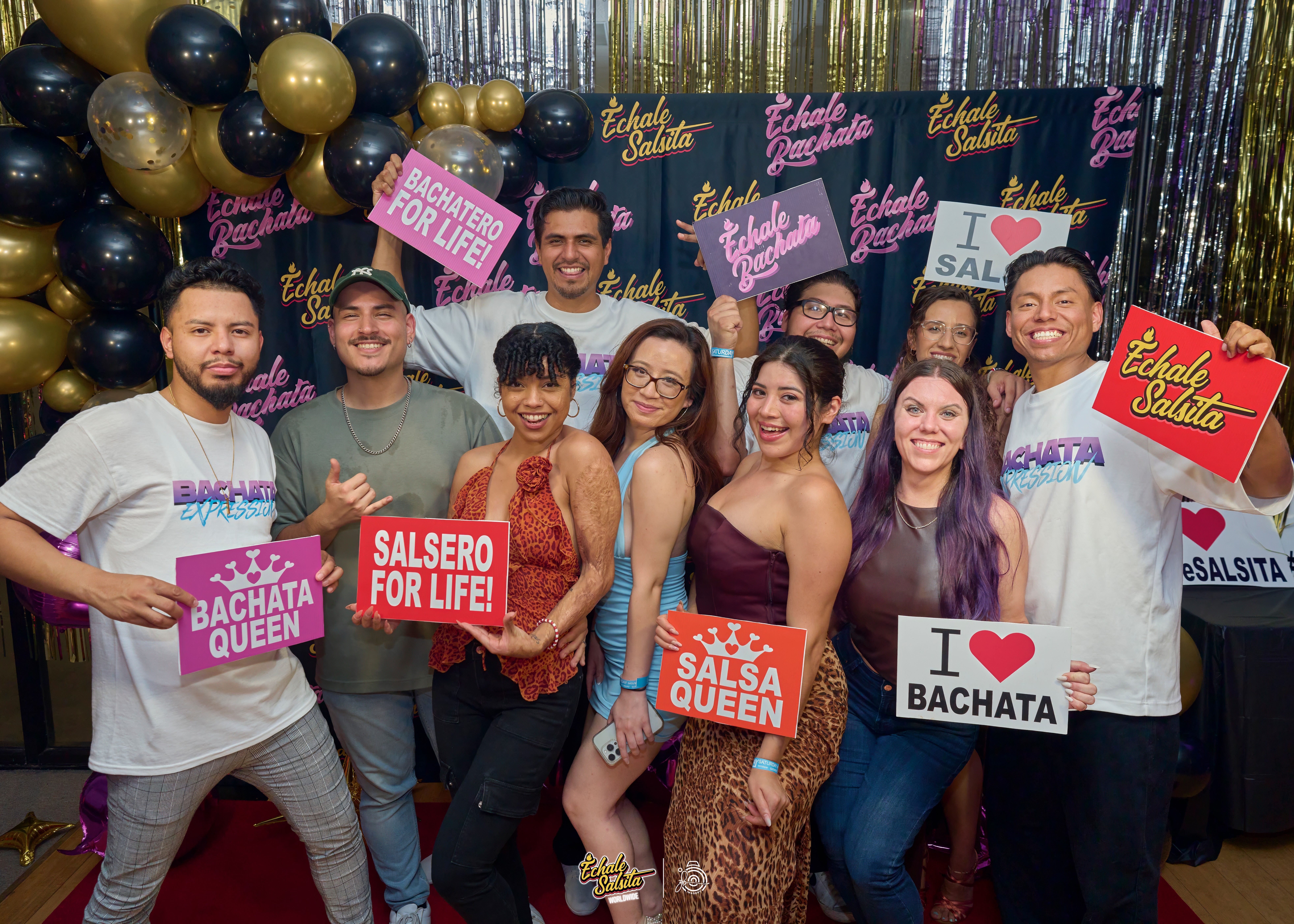 Group of people smiling and holding signs at a celebration with balloons and colorful decorations.