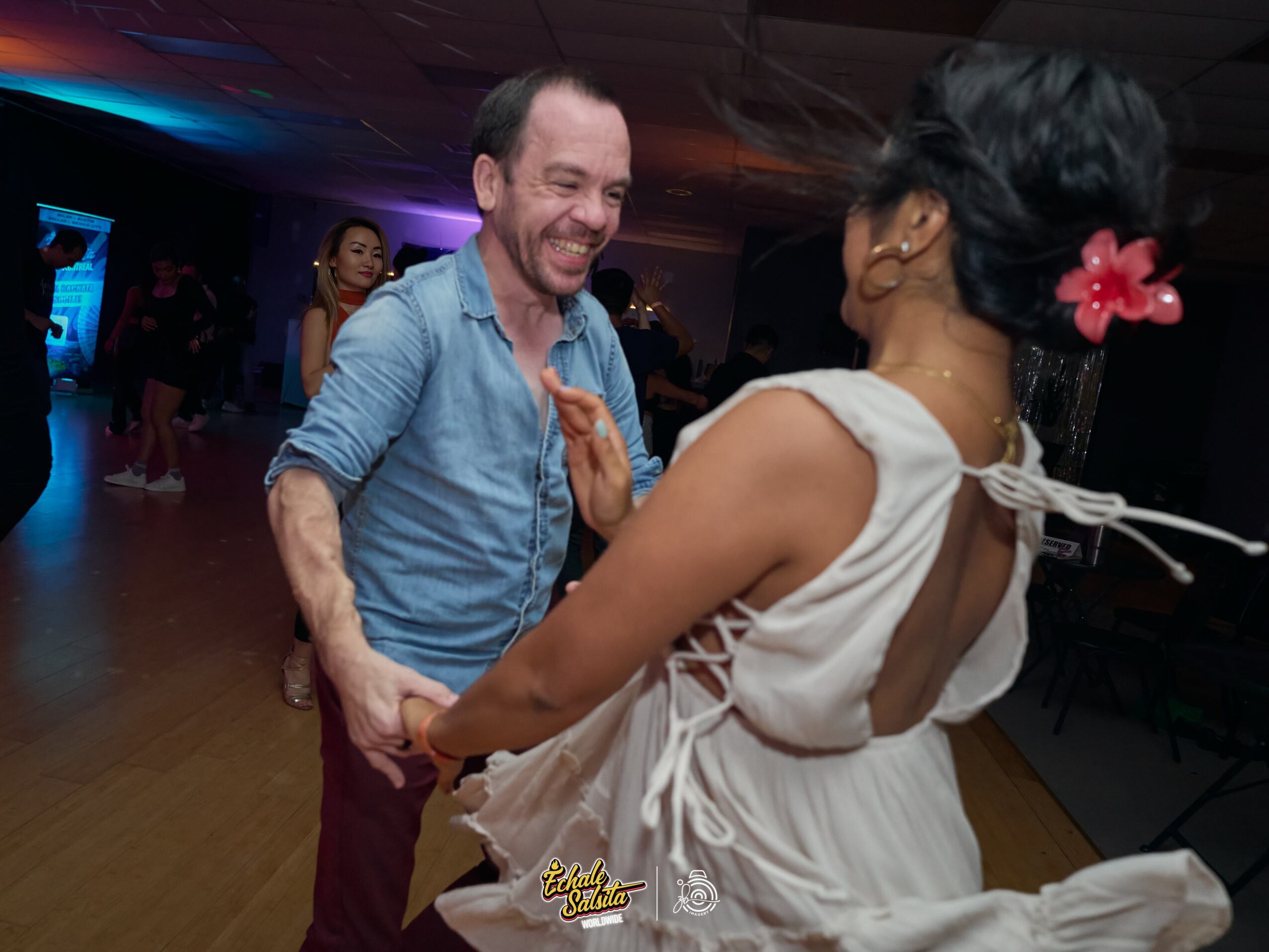 A man and woman dance together in a dimly lit room with colorful lighting, smiling and holding hands.