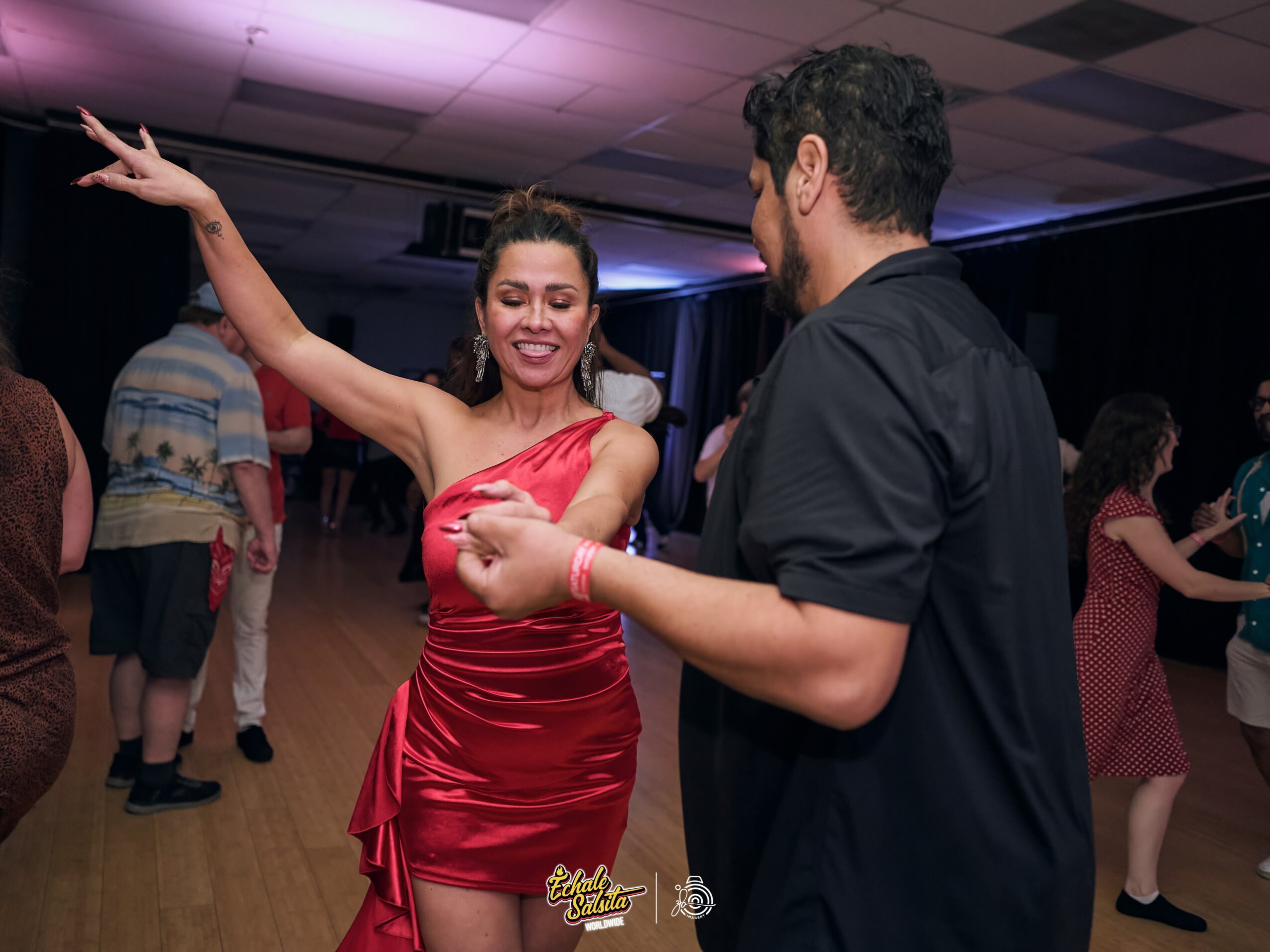 Woman in red dress dancing with a man in black shirt at a social event, other people dancing in background.