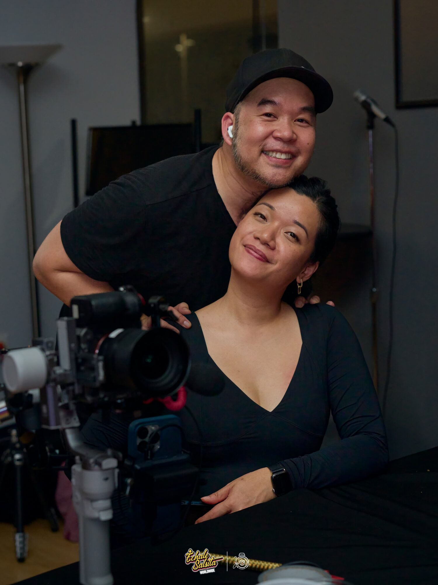 A man and woman pose together indoors, with a camera and equipment visible in the foreground.