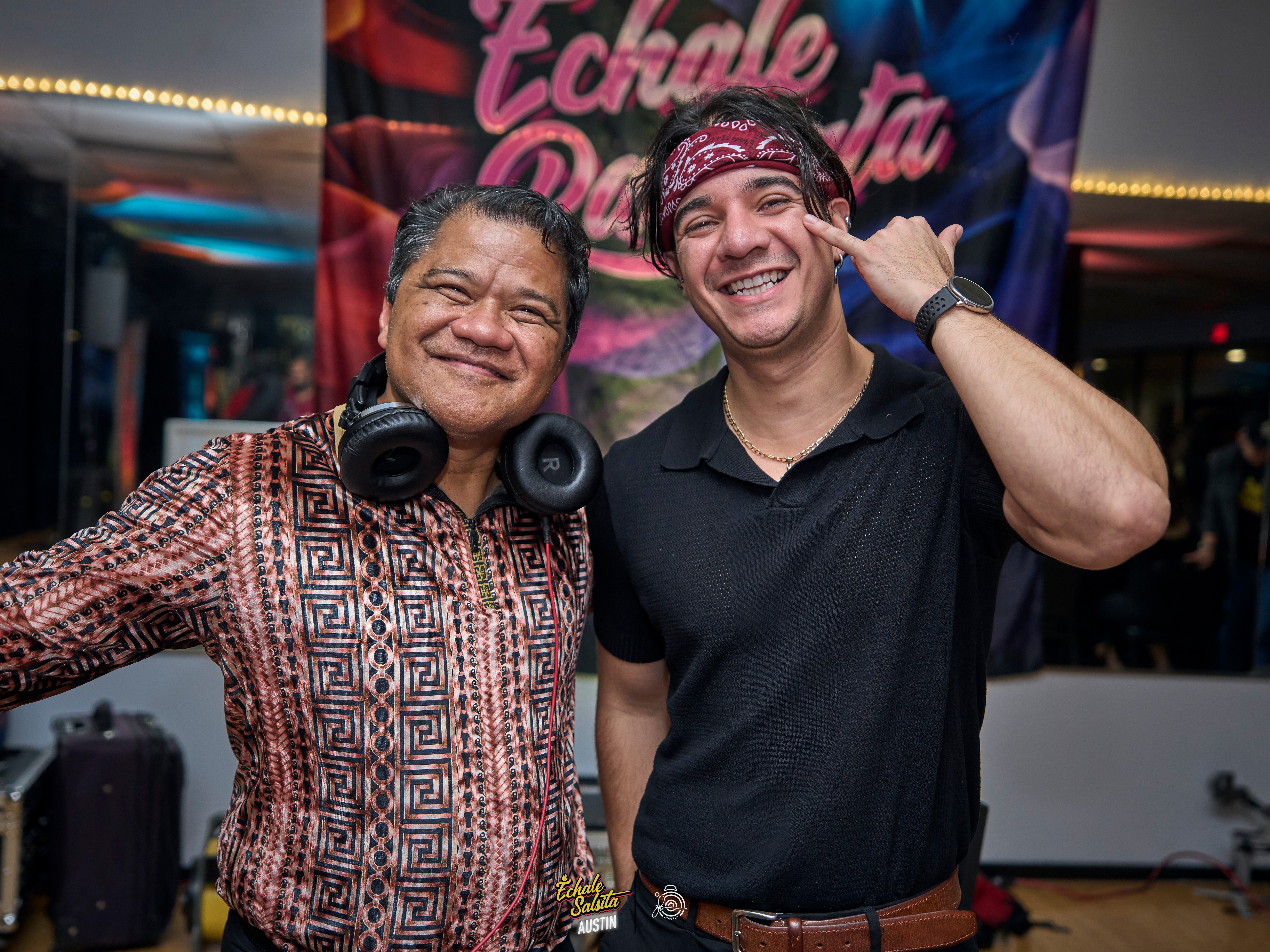 Two smiling men standing together at an event, one wearing headphones around his neck, the other making a peace sign.