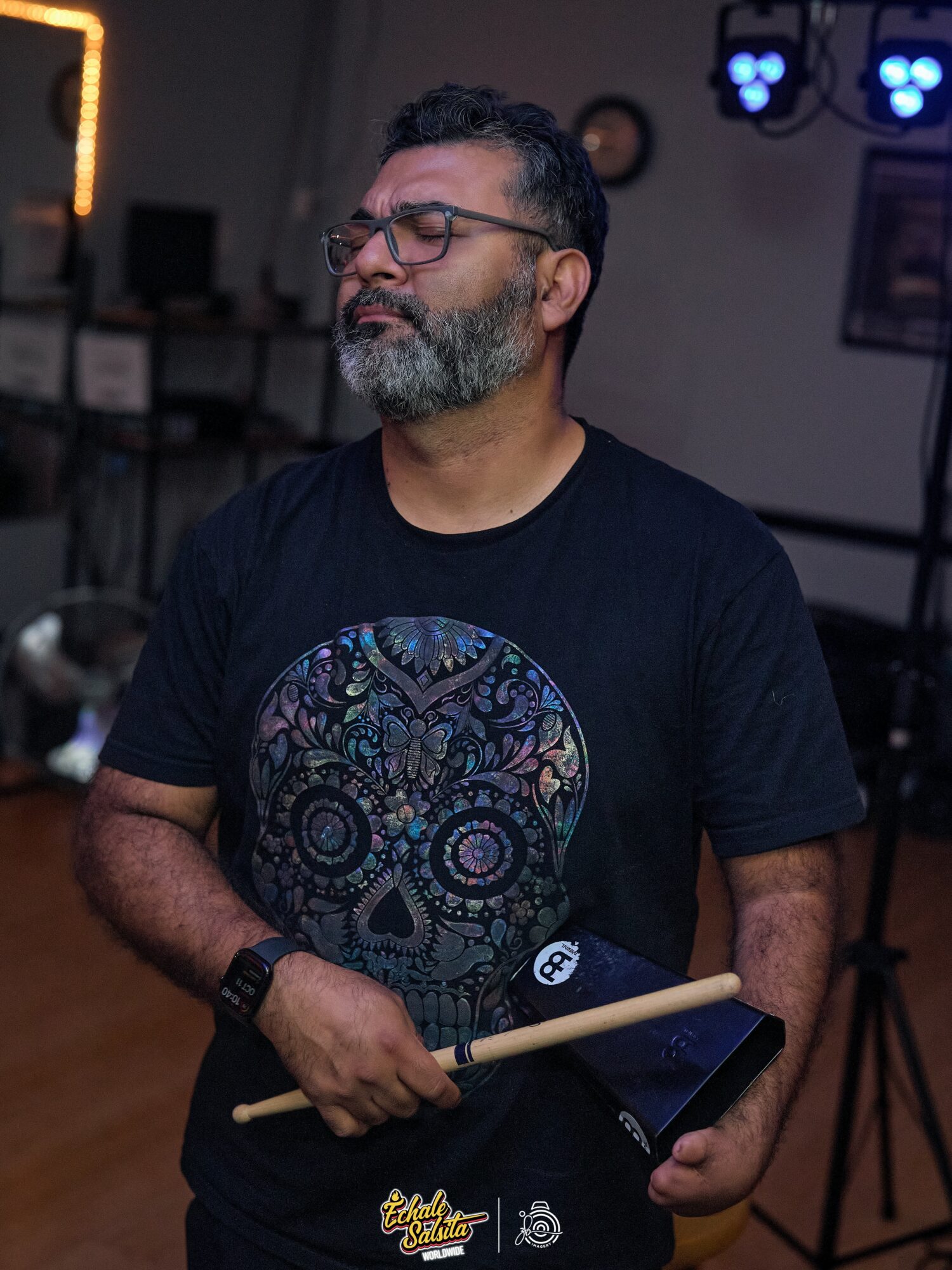 Man with glasses and a beard holding drumsticks, wearing a black T-shirt with a colorful skull design, in a dimly lit room.
