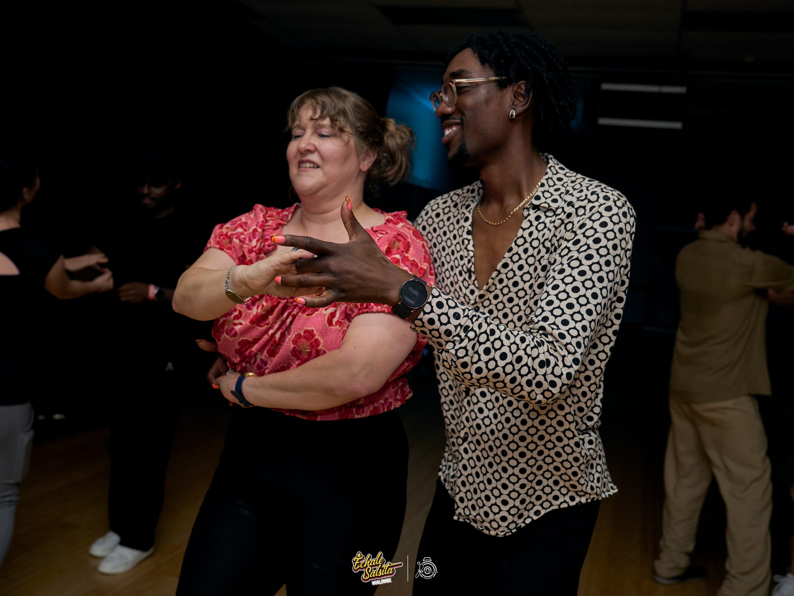 Two women dancing together, smiling, in a dark indoor setting with other people in the background.