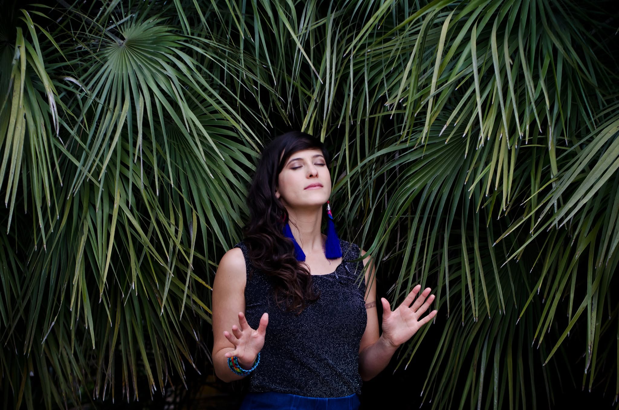 Woman with closed eyes and long dark hair stands among large green palm leaves, hands raised gently.