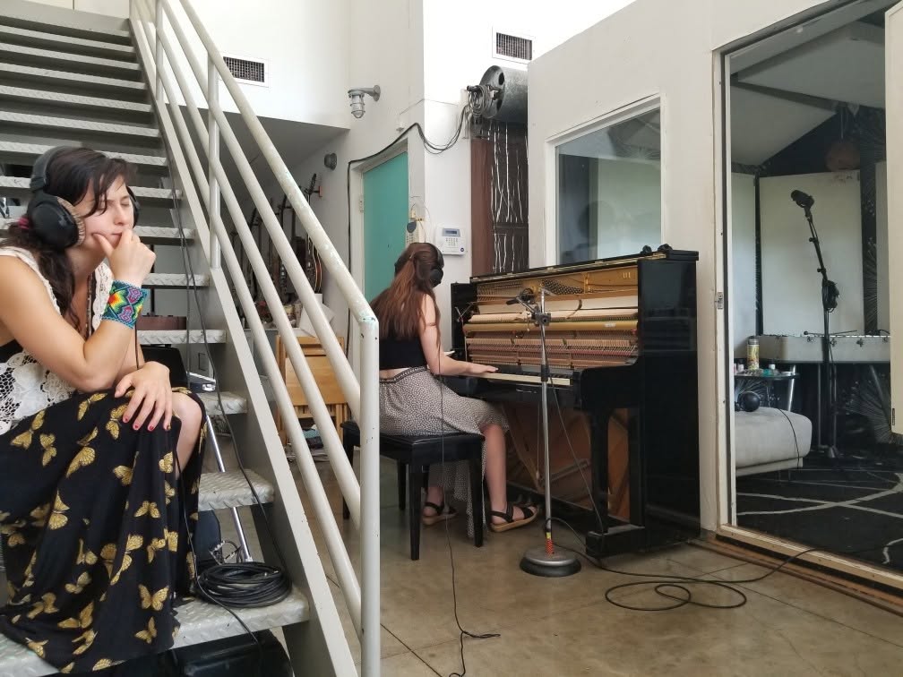 Two women in a music studio, one sitting at a piano and the other on the stairs, with recording equipment nearby.
