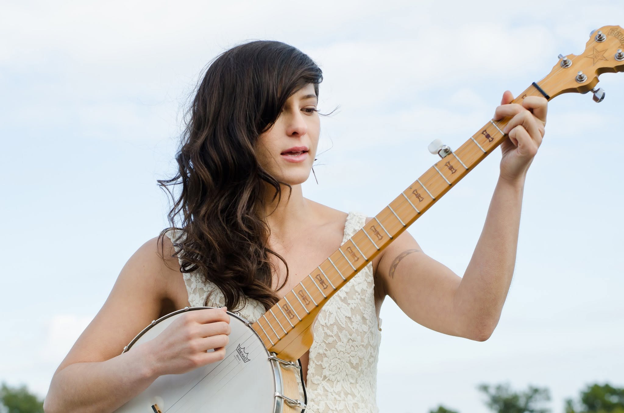 Woman with long dark hair playing a banjo outdoors against a cloudy sky, wearing a light-colored top.