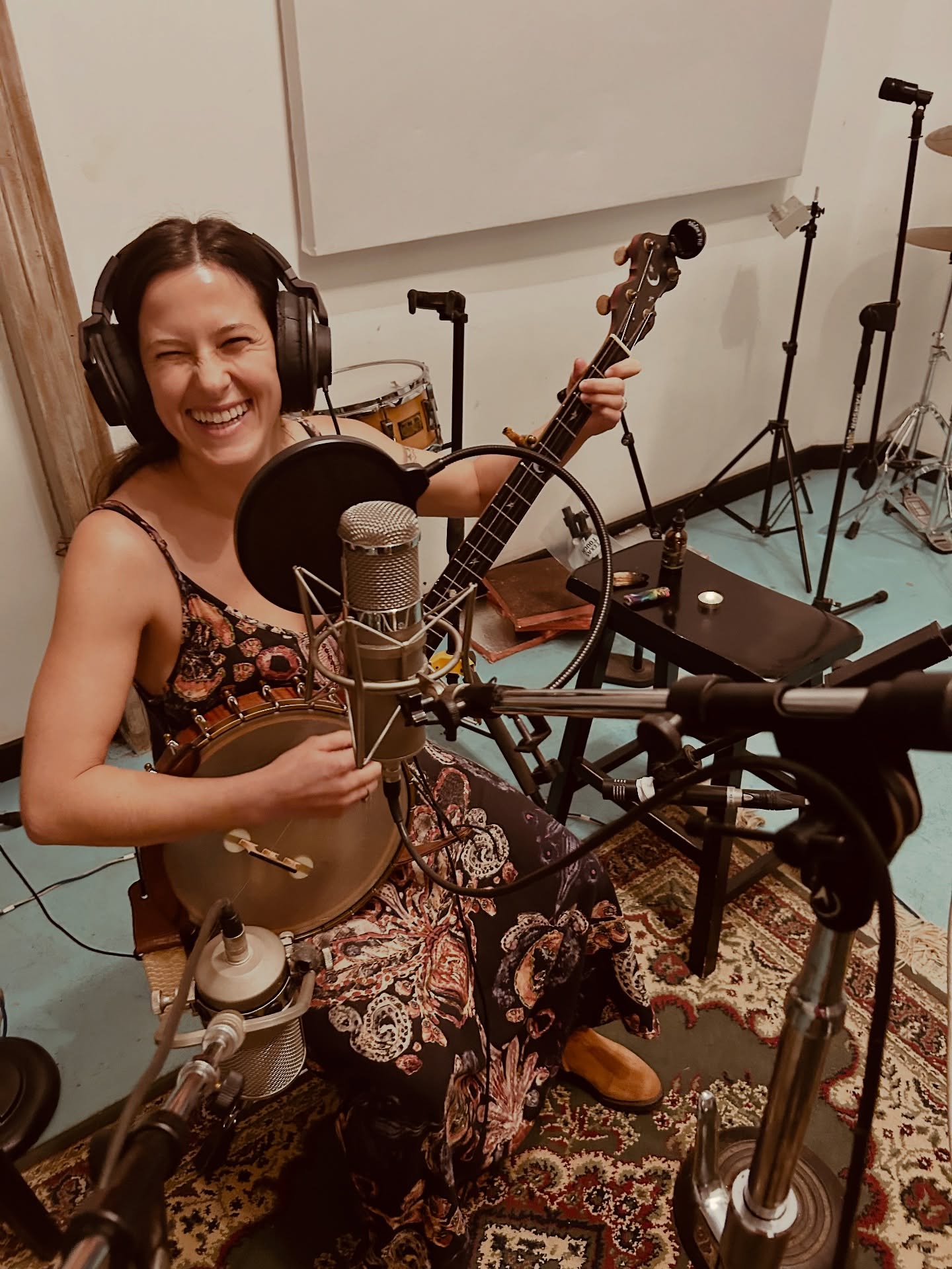 Woman smiling with headphones holding a ukulele in a recording studio, surrounded by microphones and music equipment.