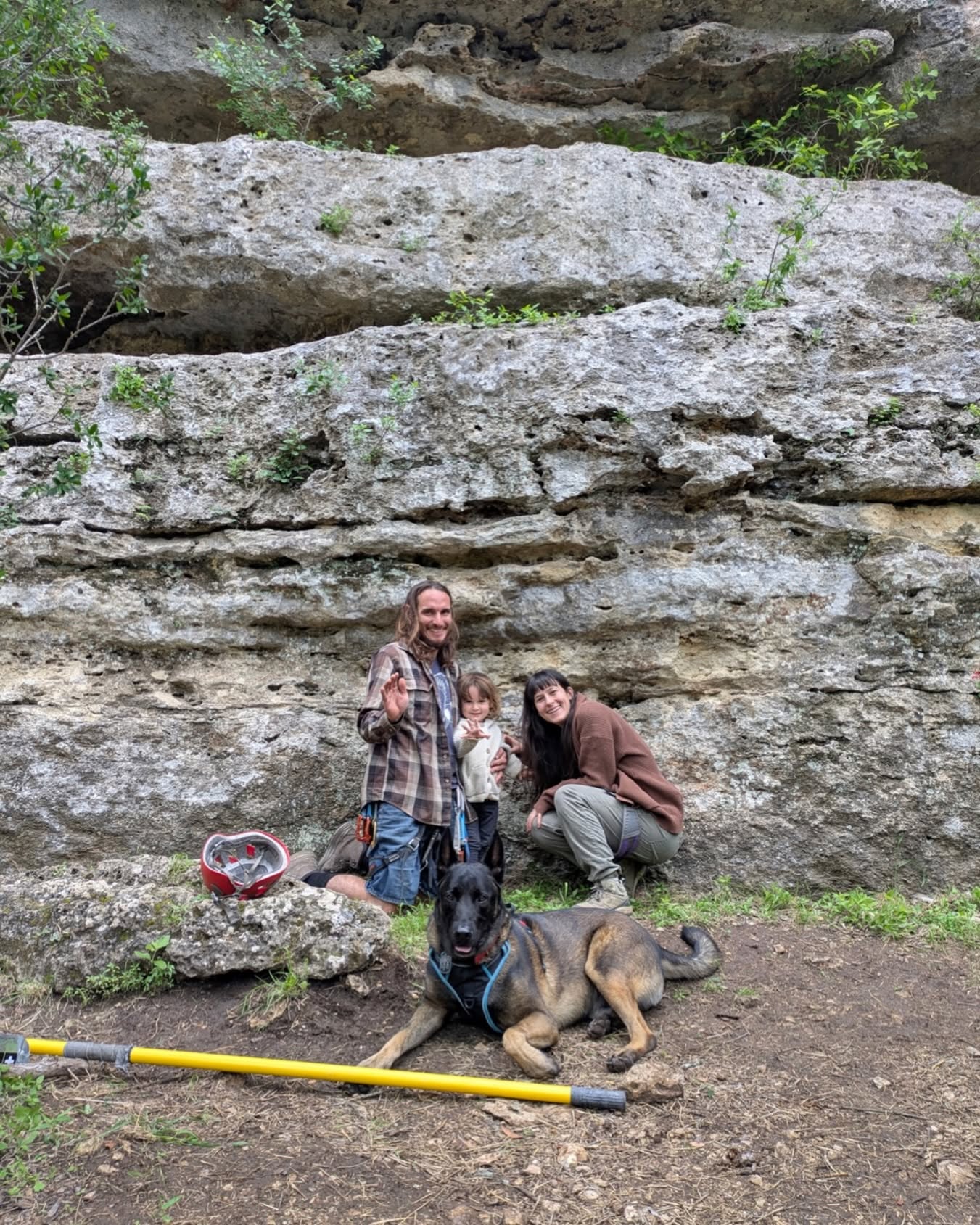 Three people and a dog in front of a large rock formation, with a yellow pole and a red and white object nearby.