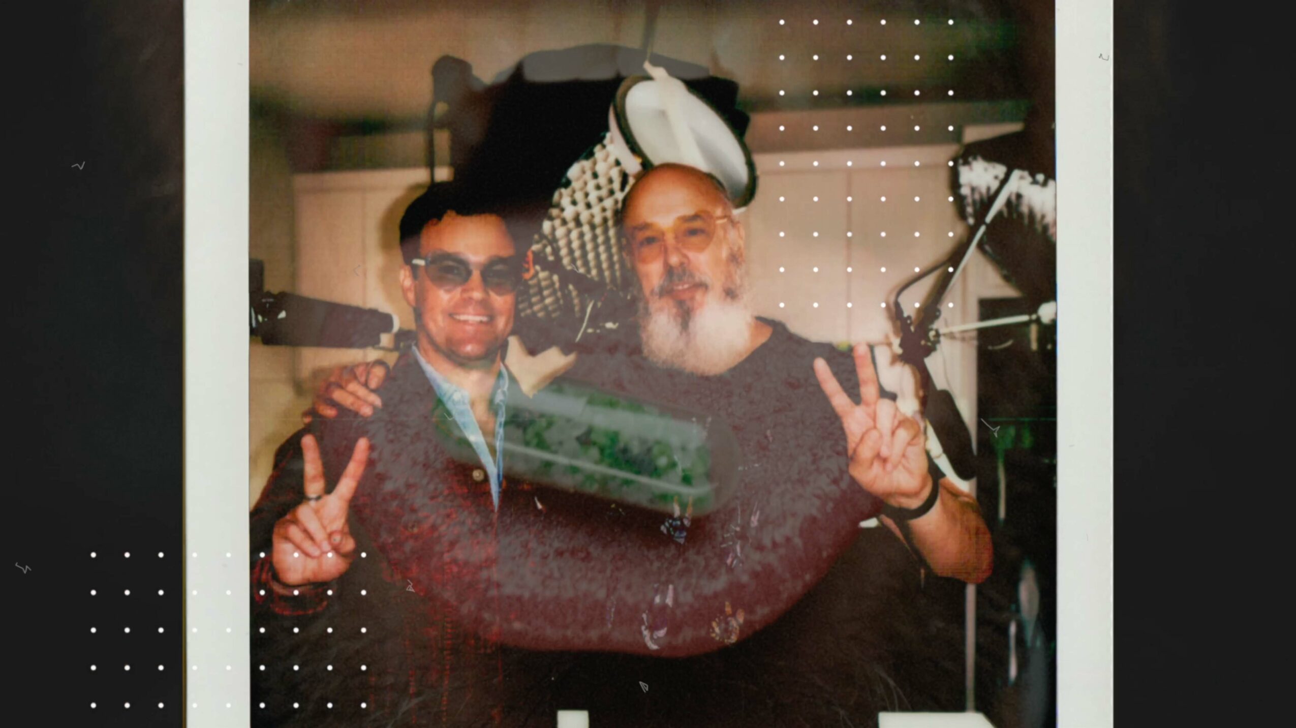 Two men standing side by side, making peace signs with their hands, in a room with various objects in the background.