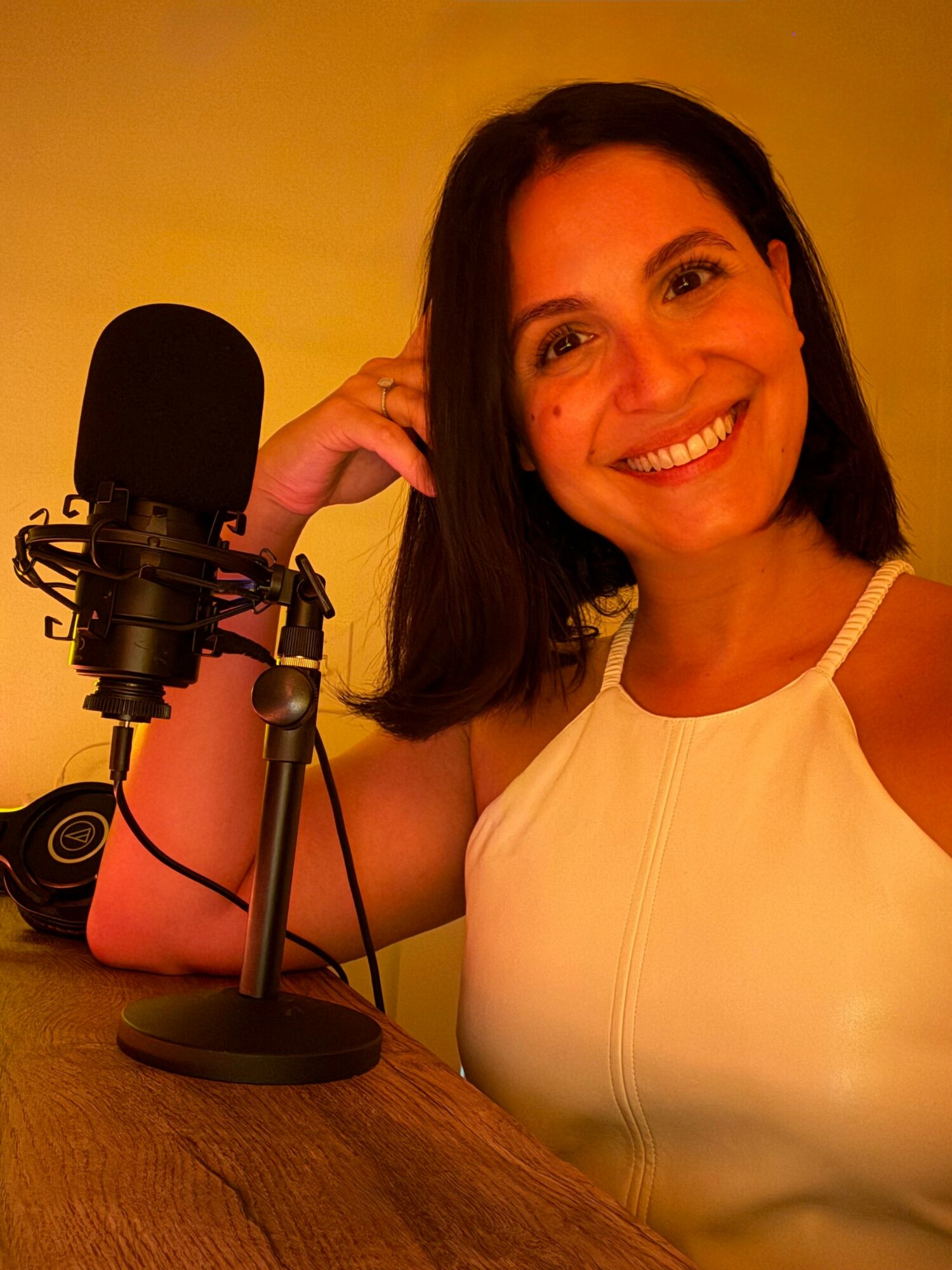 Woman smiling at microphone, wearing a white top, with a yellow background.
