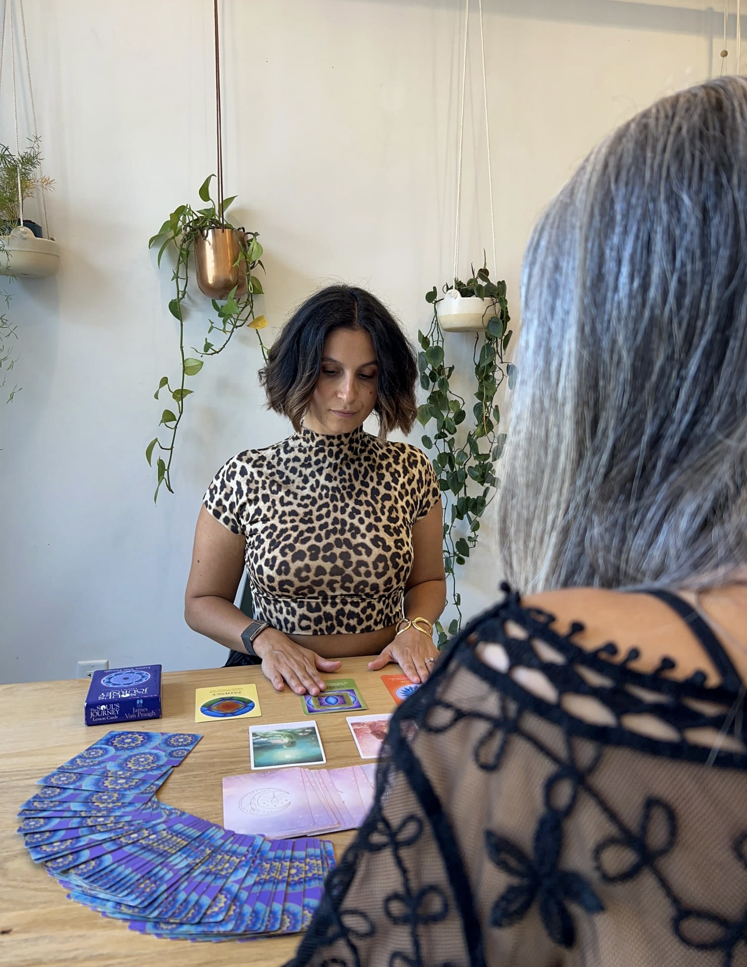 A woman with shoulder-length dark hair wearing a leopard print top stands at a table with colorful cards, in a room with hanging plants.