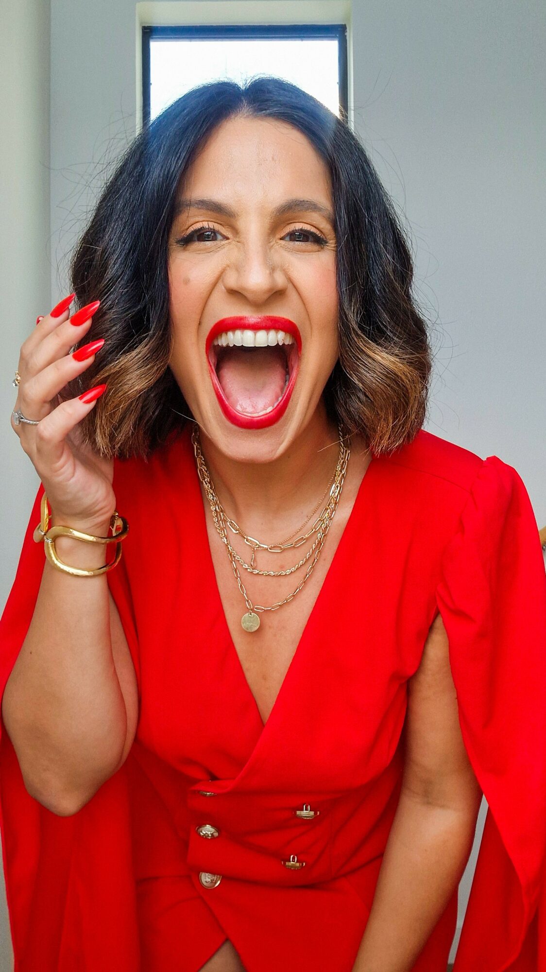 Woman with shoulder-length dark hair, laughing with mouth open, wearing a red dress and gold jewelry, in a room with a window