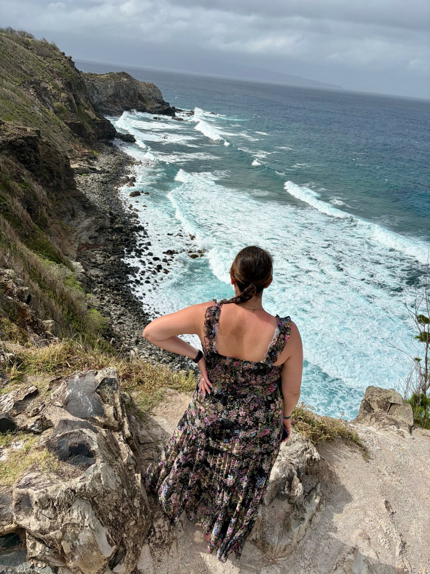 Woman in a floral dress sitting on rocks overlooking a rocky coastline with waves crashing below.