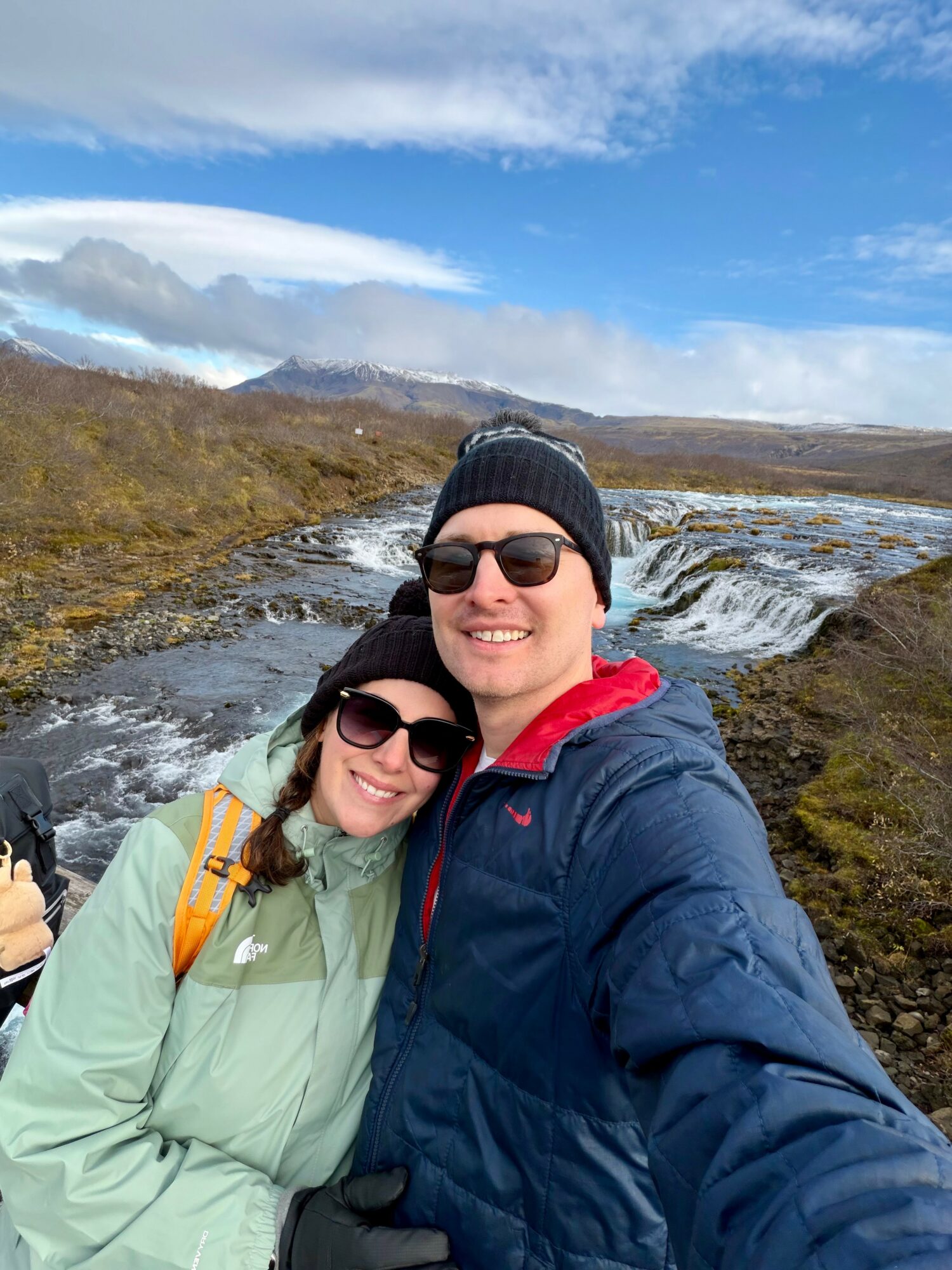 A smiling couple wearing sunglasses and hats outdoors near a river with mountains and blue sky in the background.