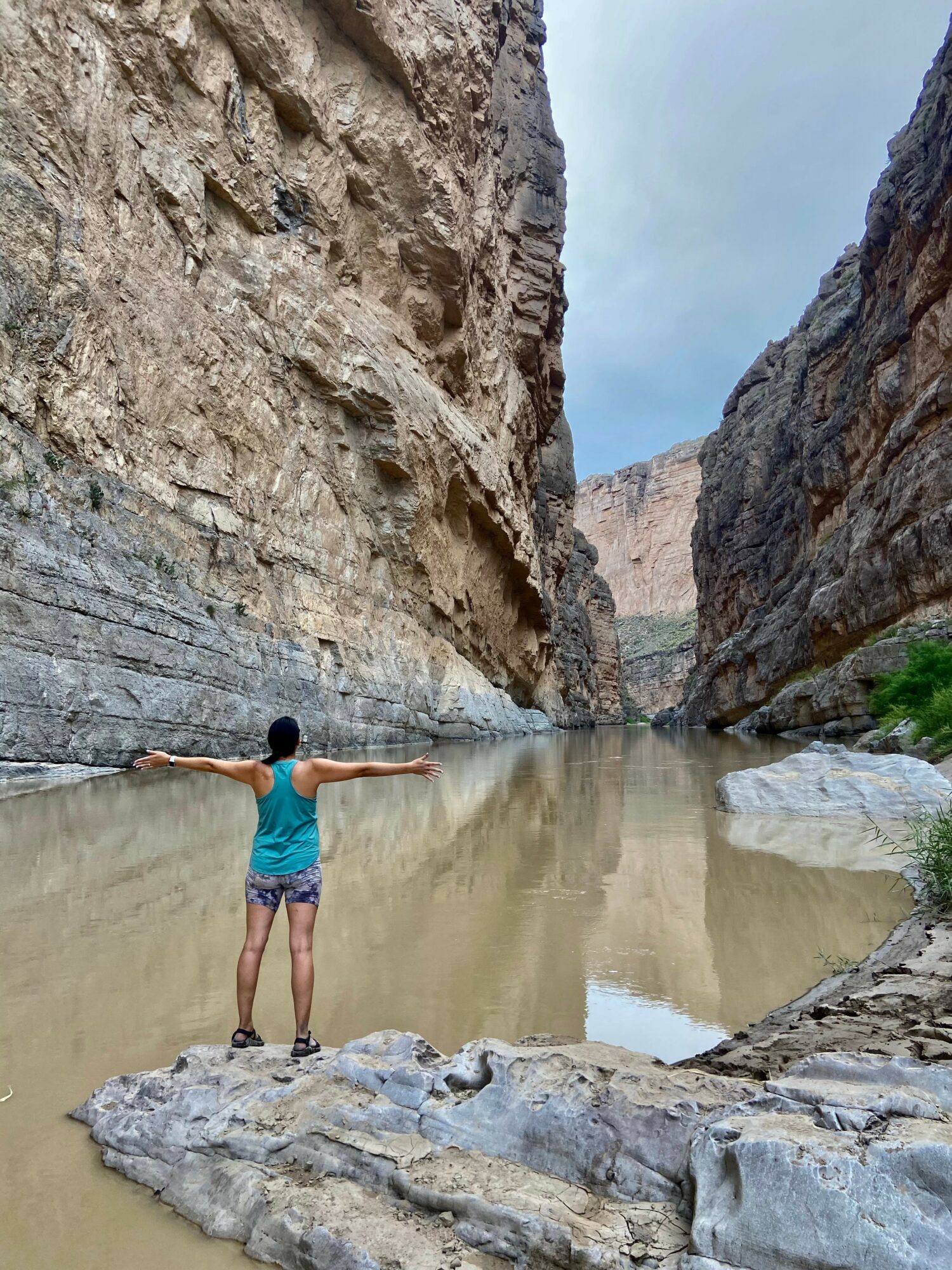Person standing with arms outstretched in a narrow canyon with a river, high rocky walls, and a cloudy sky.