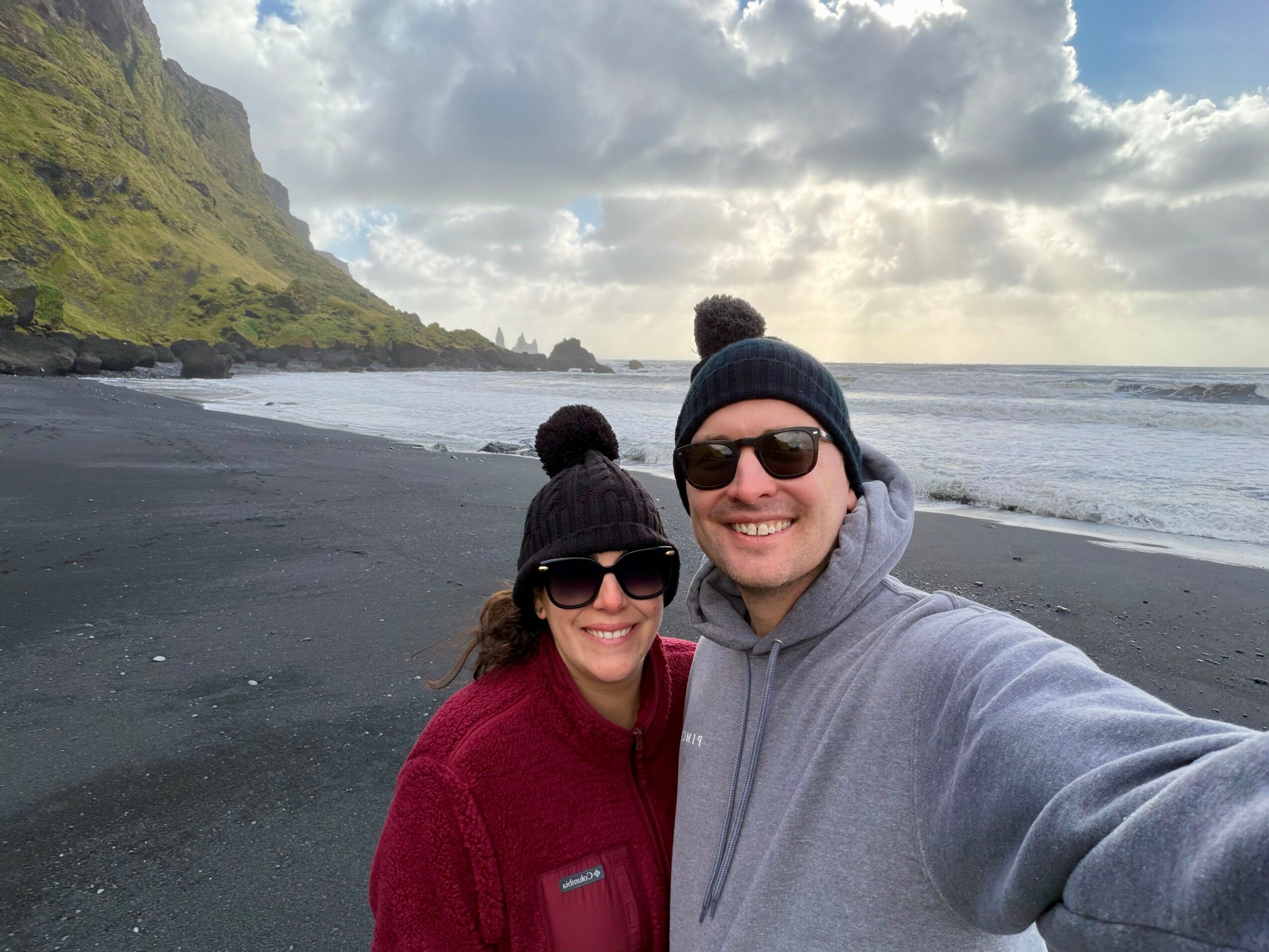 Two people wearing winter clothing and sunglasses taking a selfie on a black sand beach with cliffs and cloudy sky in the background.