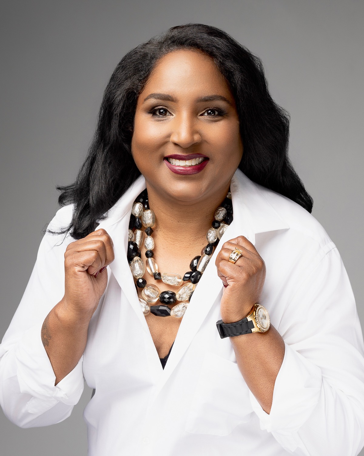 Woman with long dark hair wearing a white blazer, layered necklaces, and a watch, smiling confidently against a gray background.