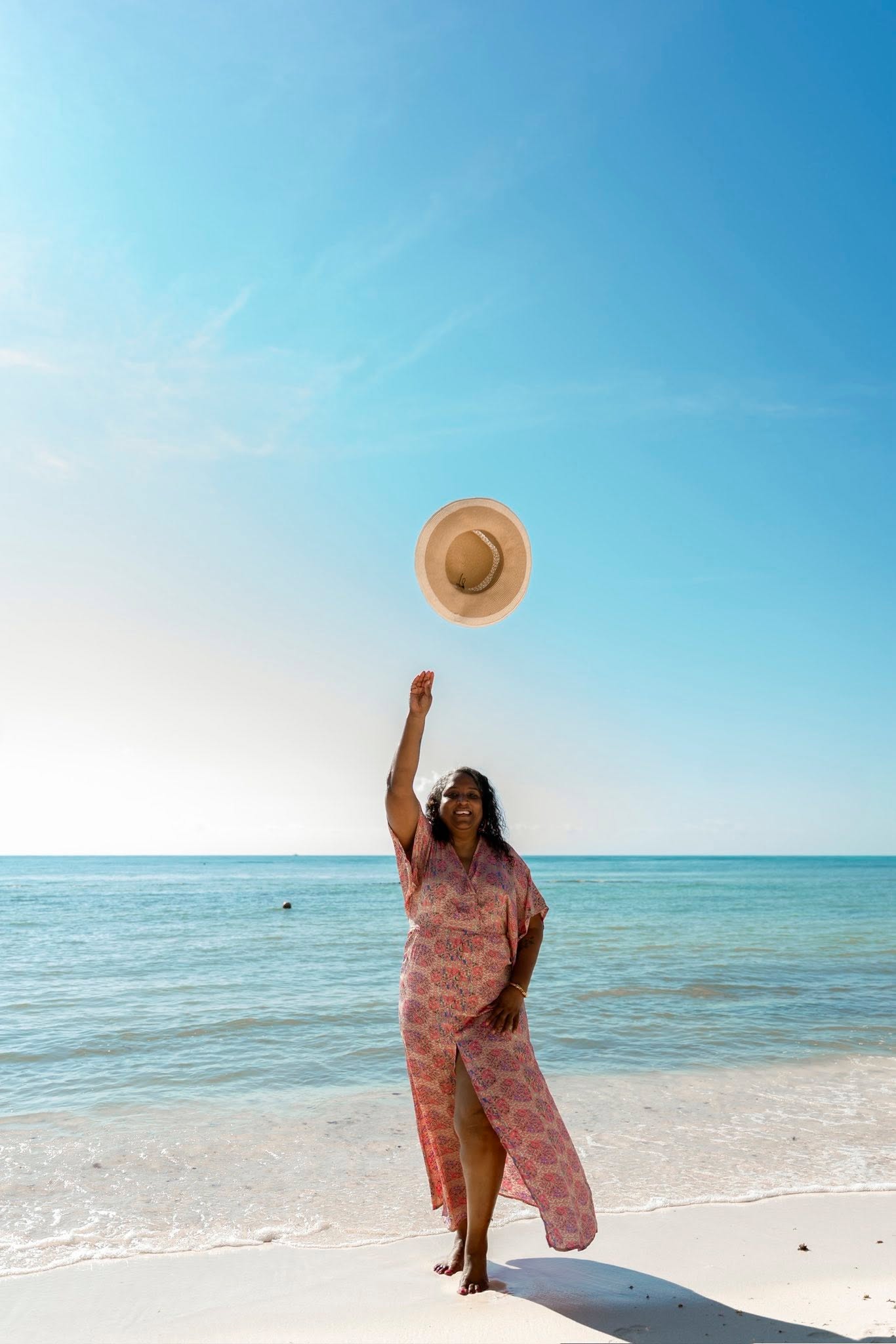 Woman in pink dress throws hat into clear blue sky at beach, ocean in background, sunny day.