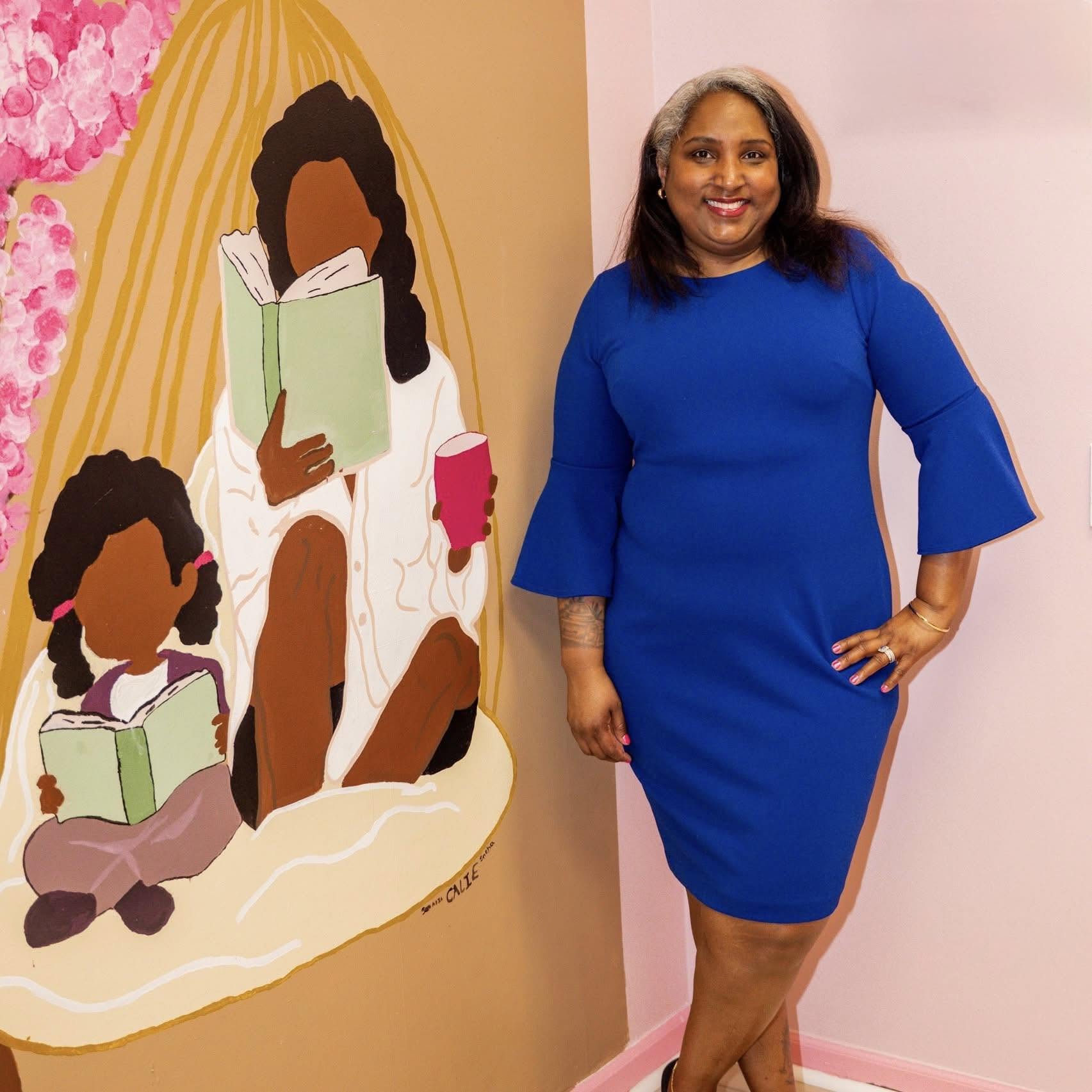 Woman in blue dress standing next to a colorful mural of two people reading books, one adult and one child, on a pink wall.