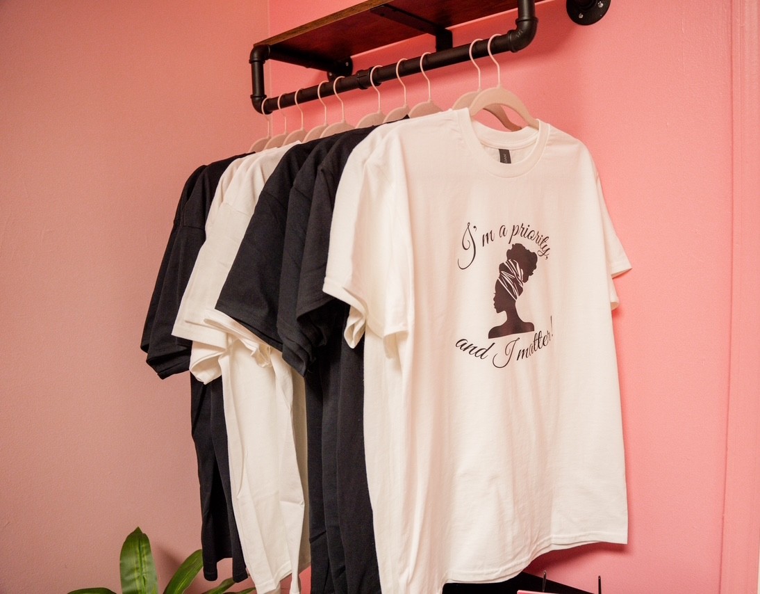 White T-shirt with black text and graphic hanging on a rack, with black and white shirts behind, pink wall background
