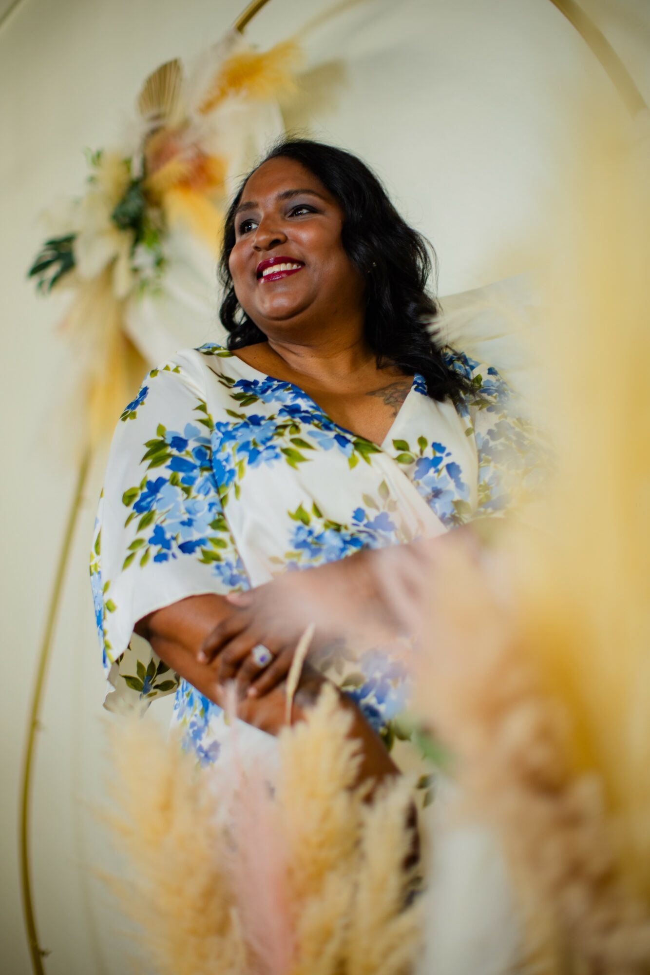 Woman with dark hair smiling, wearing a floral dress, standing in front of a decorated background with flowers.