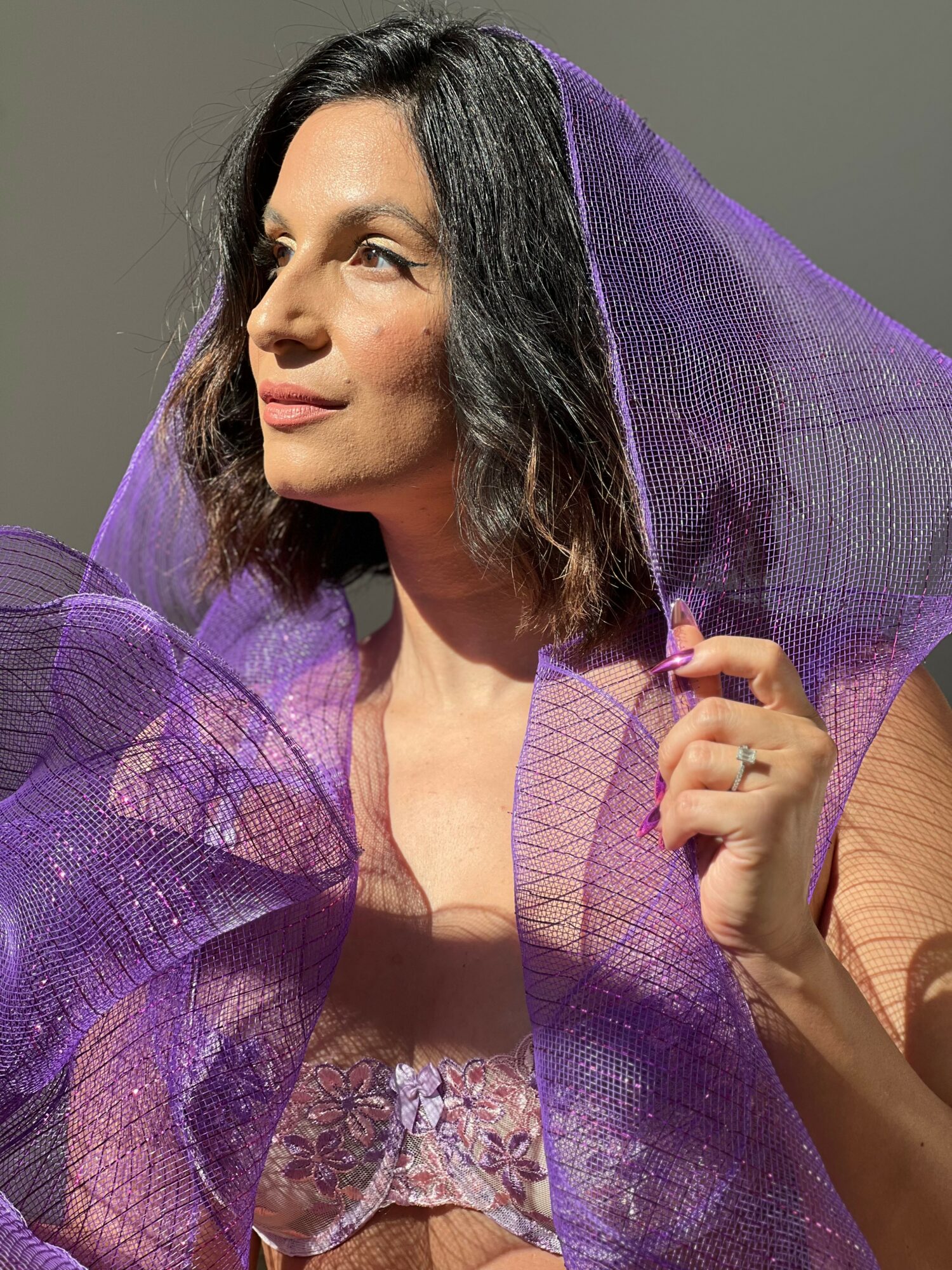 Woman with dark hair holding purple sheer fabric, looking to the side, gray background.