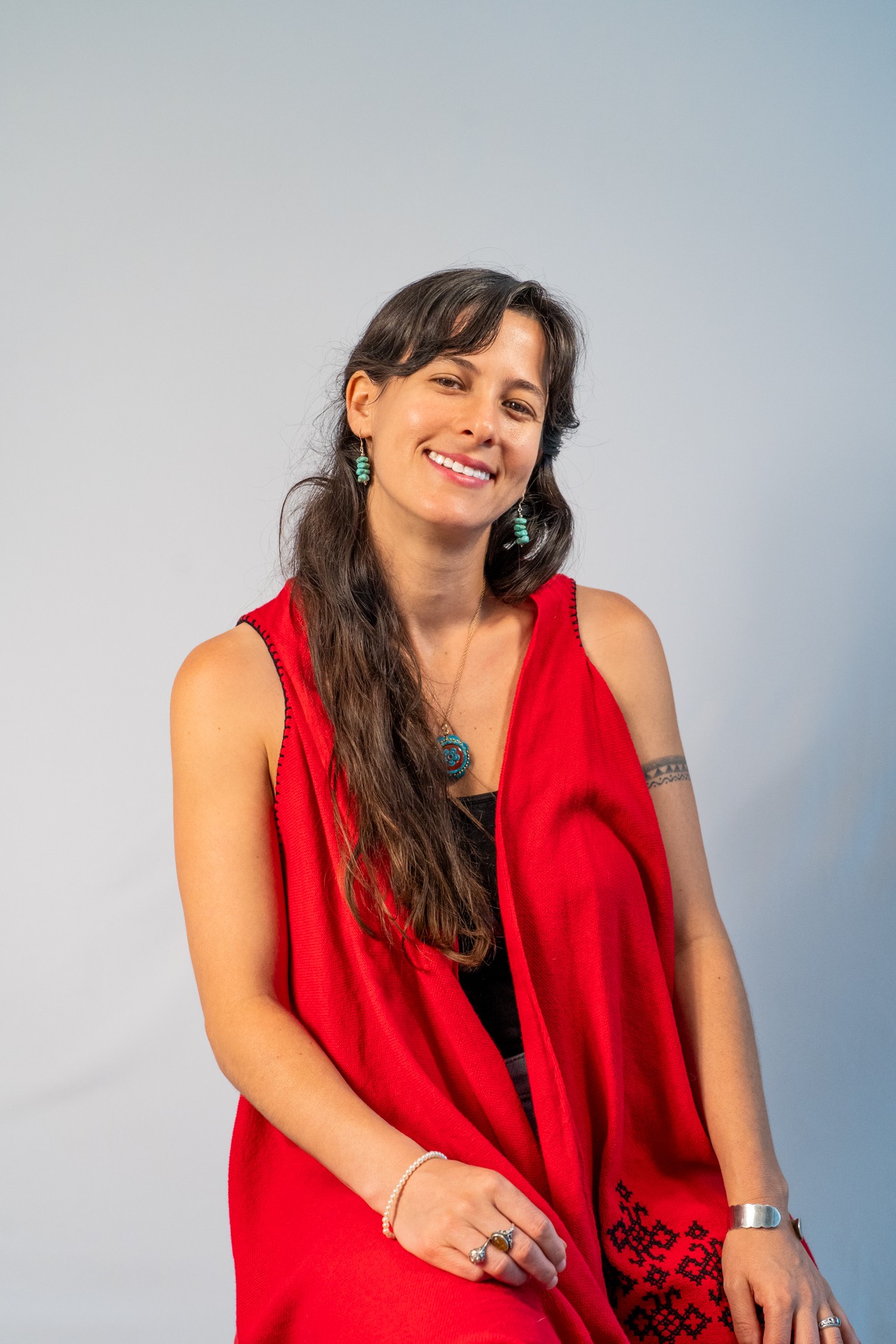 Young woman with long dark hair smiling, wearing a red sleeveless top and jewelry, against a plain background.