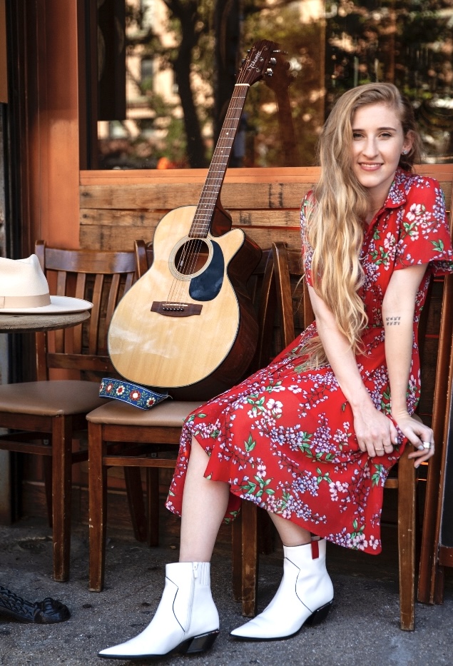 Young woman with long blonde hair in a red floral dress and white boots sitting on a wooden chair outdoors, with an acoustic guitar beside her.