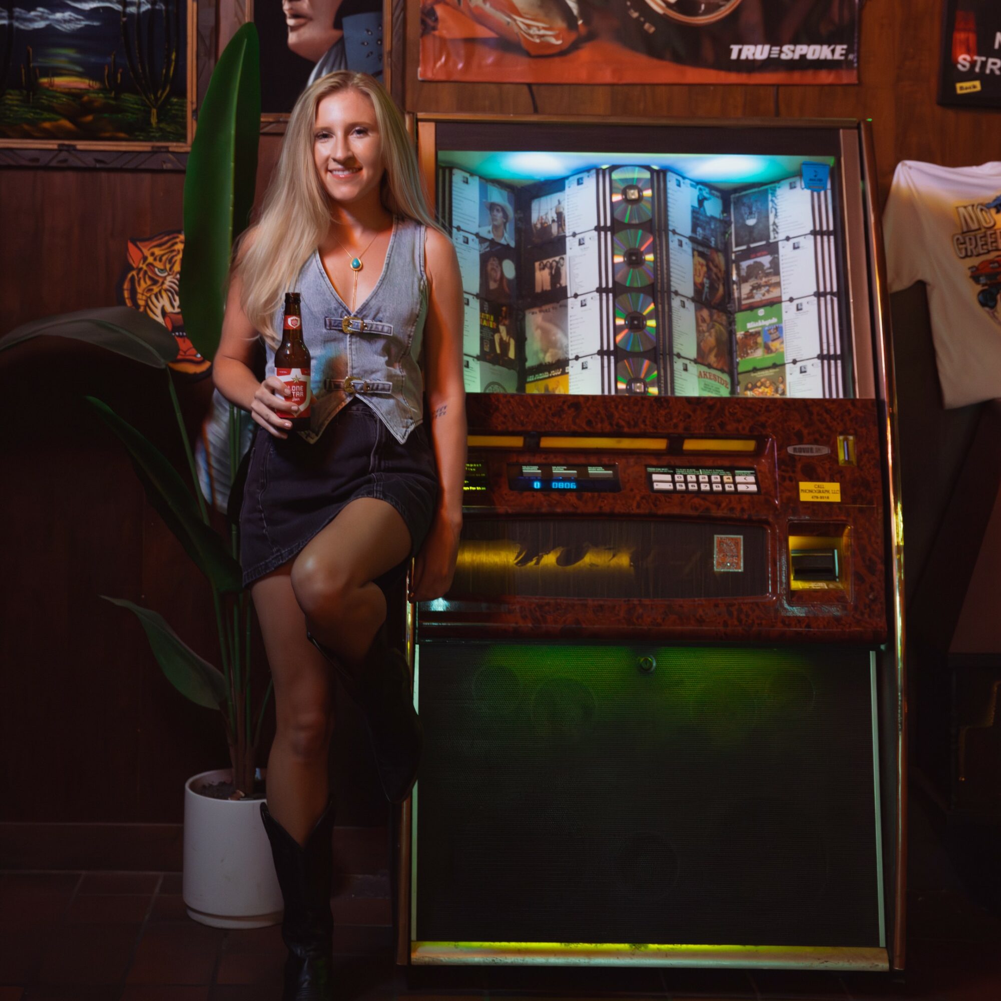 Woman with long blonde hair holding a drink, standing next to a vintage arcade game machine in a cozy room.