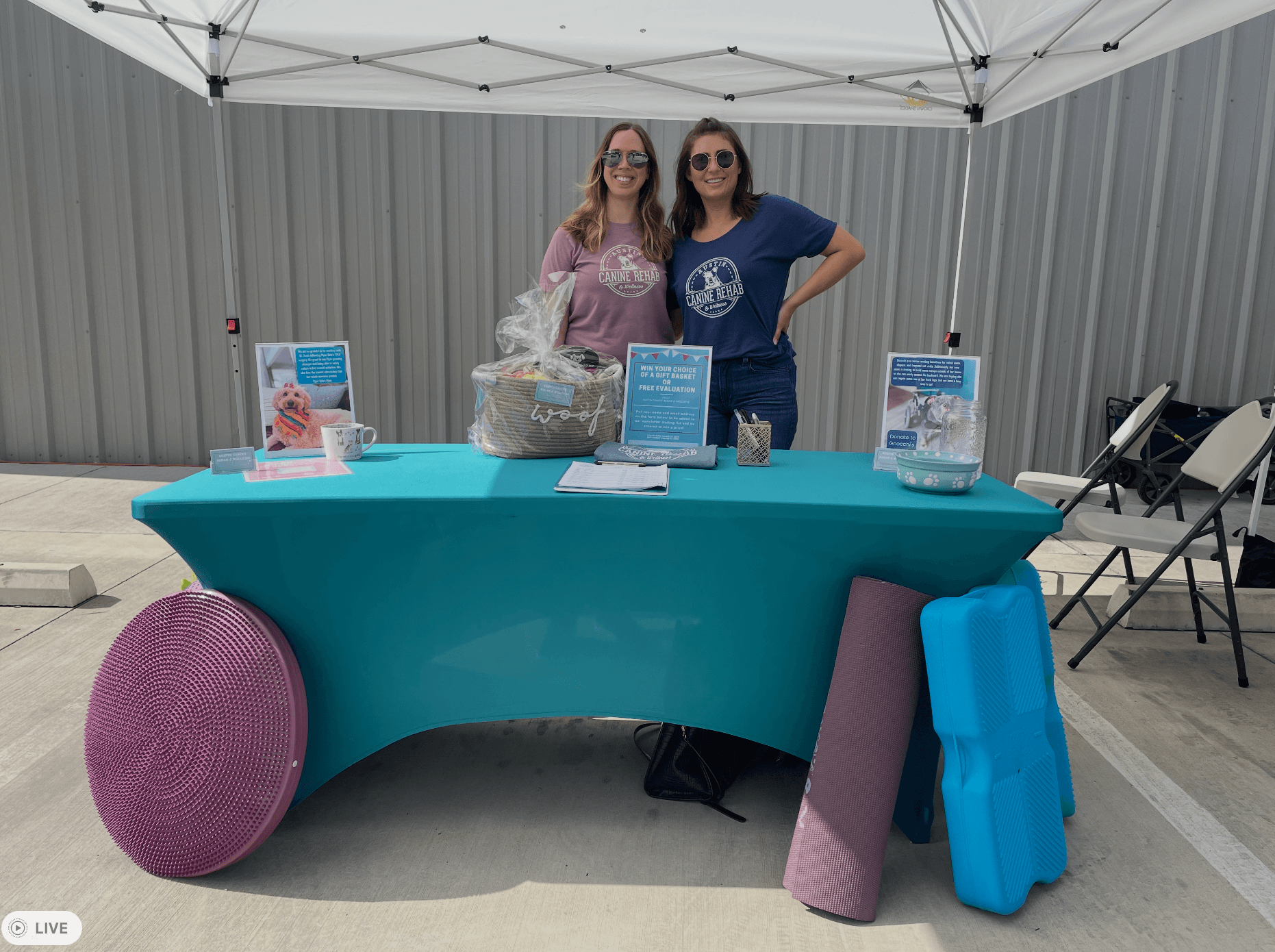 Two women stand behind a table under a white canopy, with informational signs and items displayed, in an outdoor setting.