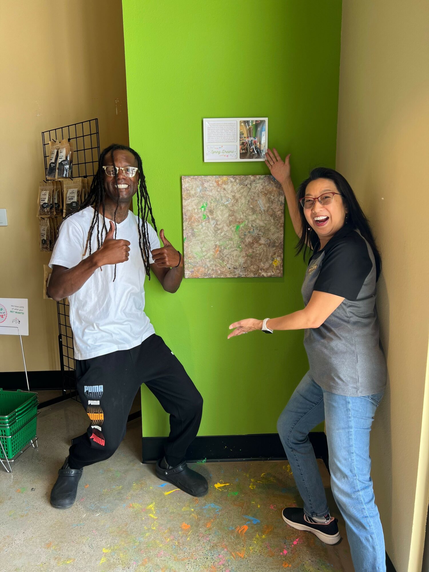 Two women posing indoors, one standing and leaning against a green wall, the other sitting on the floor with a colorful carpet.