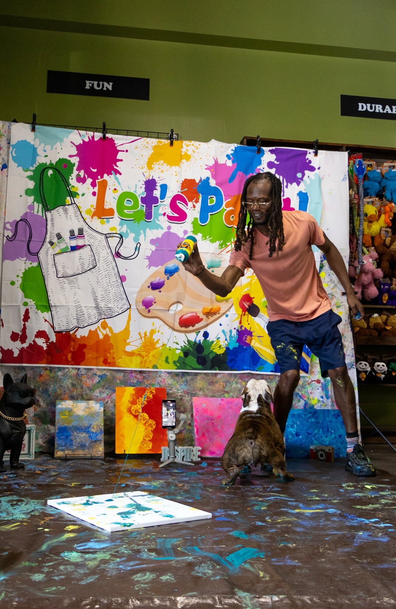 Person with long hair and pink shirt painting a colorful mural with a dog nearby, store shelves in background.