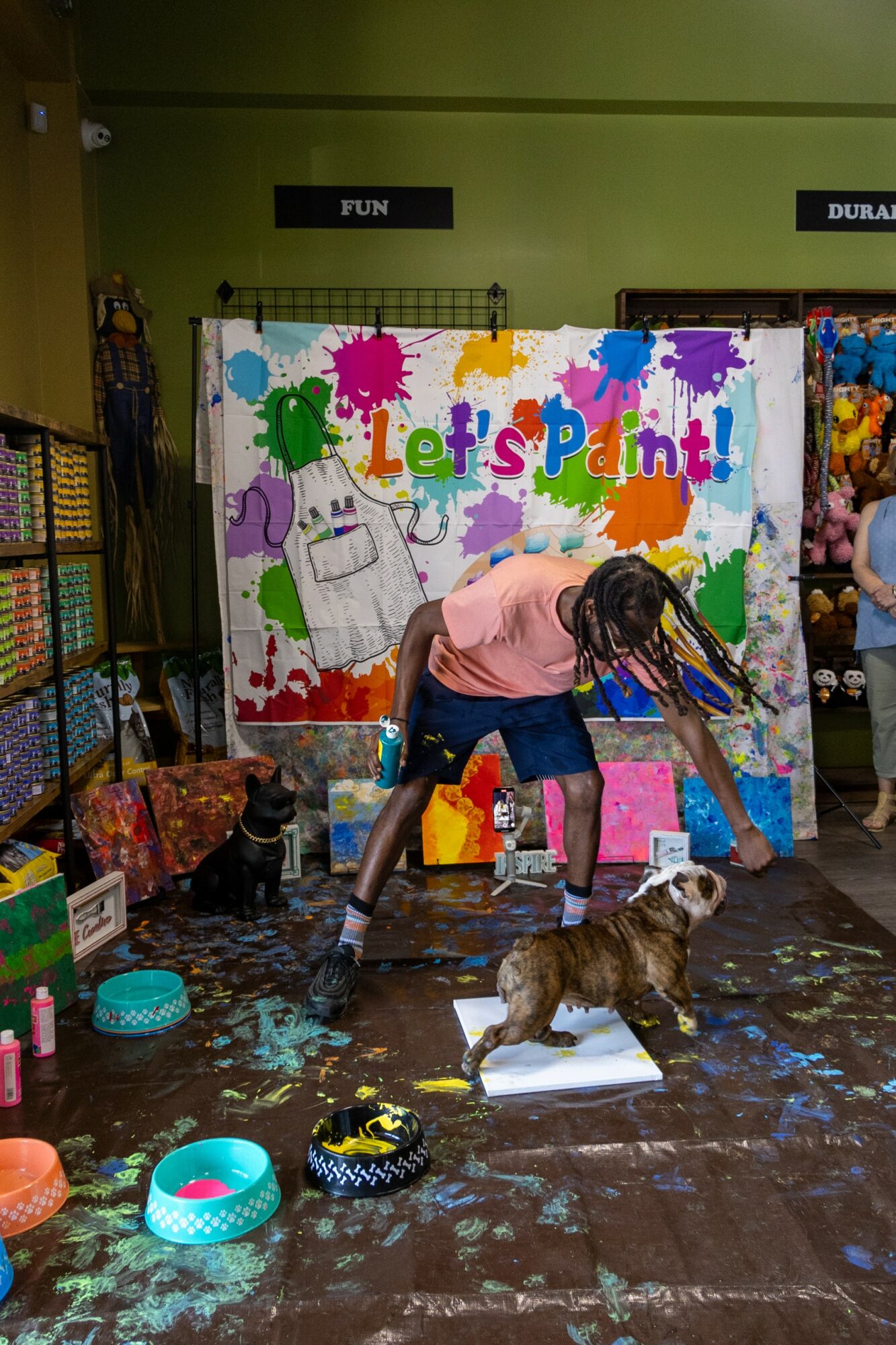 Person with long hair and pink shirt painting on a colorful wall with a dog nearby, in a room with art supplies.