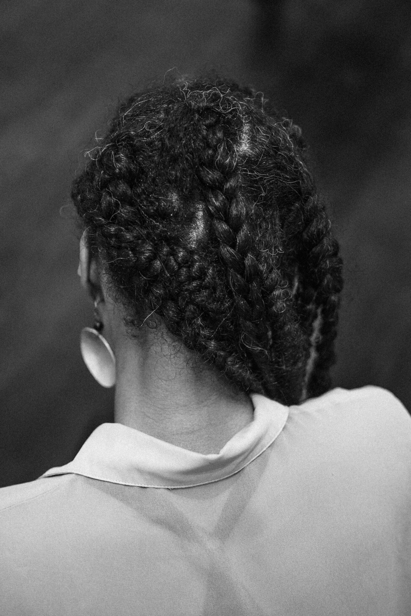 Back of a person's head with textured, braided hairstyle, wearing earrings and a collared shirt, in black and white.