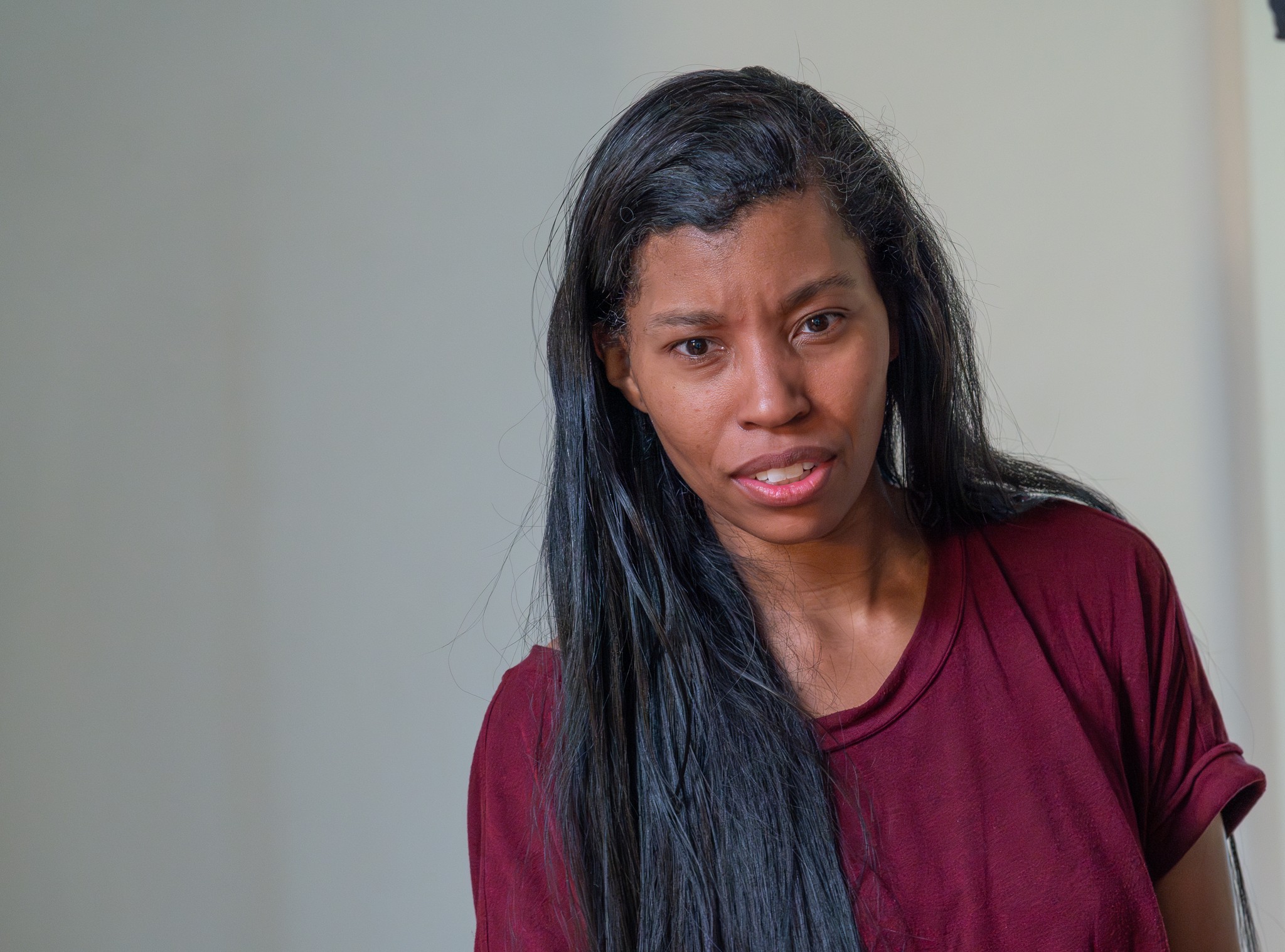 Woman with long black hair wearing a maroon shirt, looking confused or concerned, standing indoors.