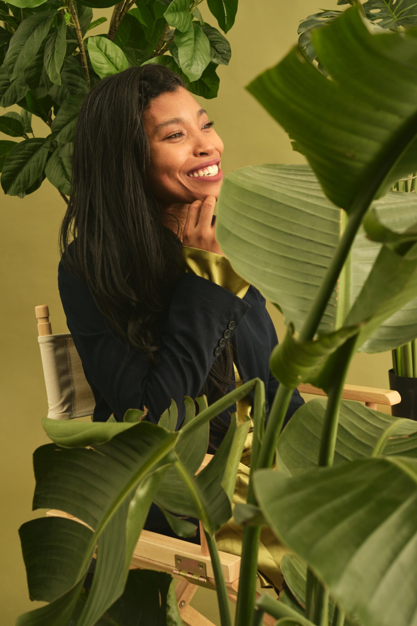 Smiling woman with long dark hair sitting among large green plants, touching her face gently, in a well-lit room.