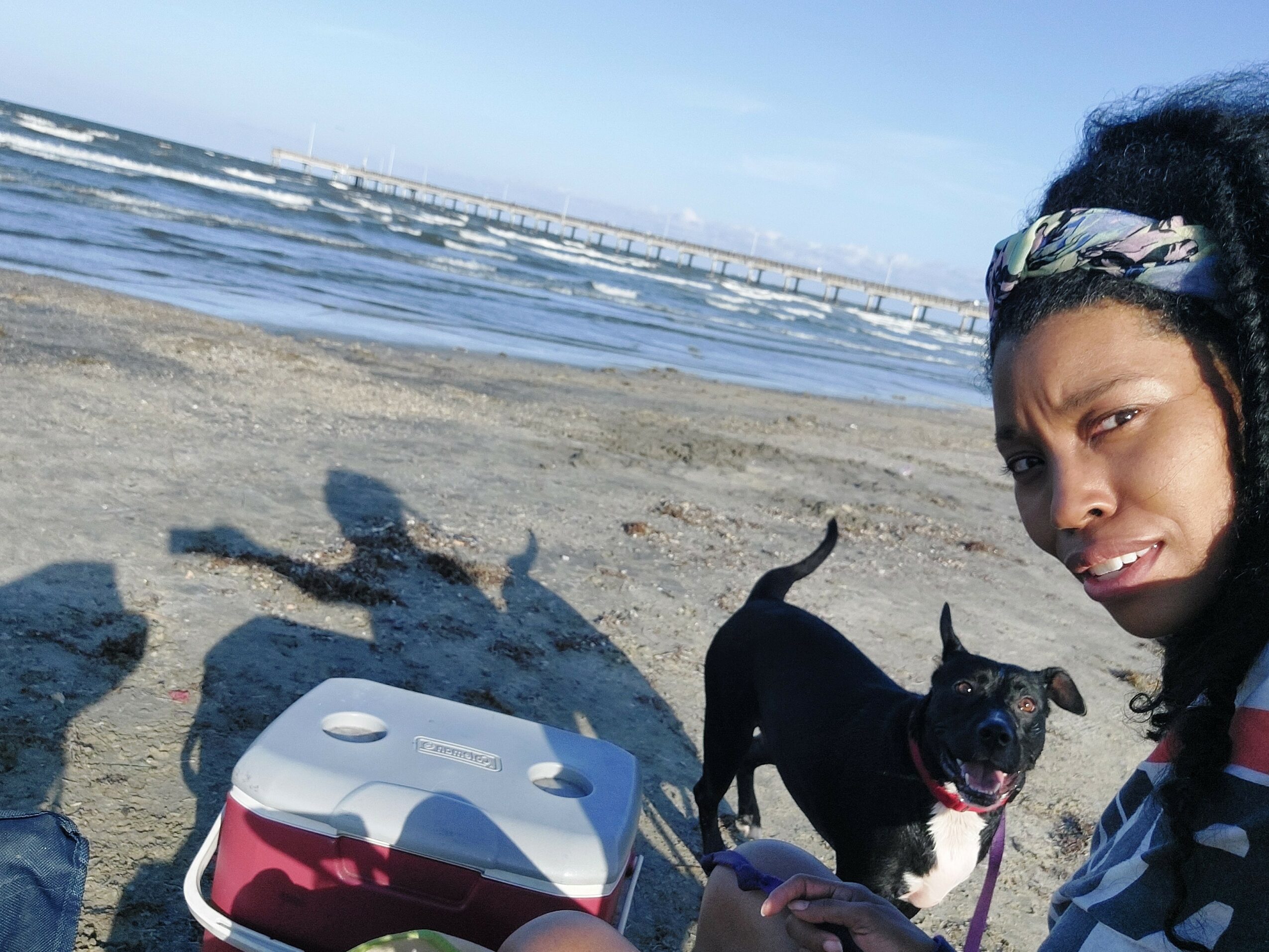 Person with dark hair and headband taking selfie on beach with black dog, ocean, and pier in background.