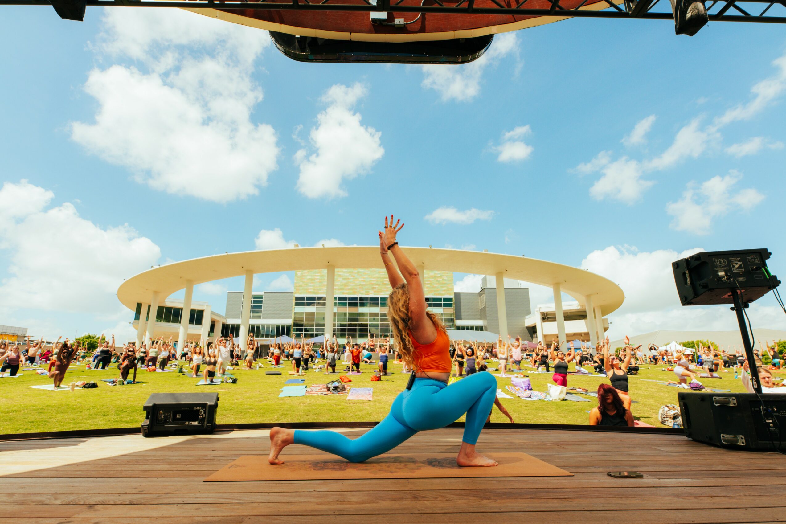 Person performing yoga pose on stage outdoors with a large circular building and crowd in background, blue sky with clouds.