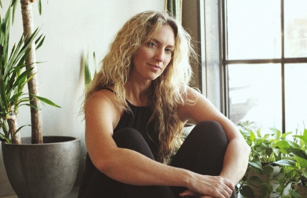 Woman with long curly hair sitting by a window, surrounded by plants, looking at the camera.