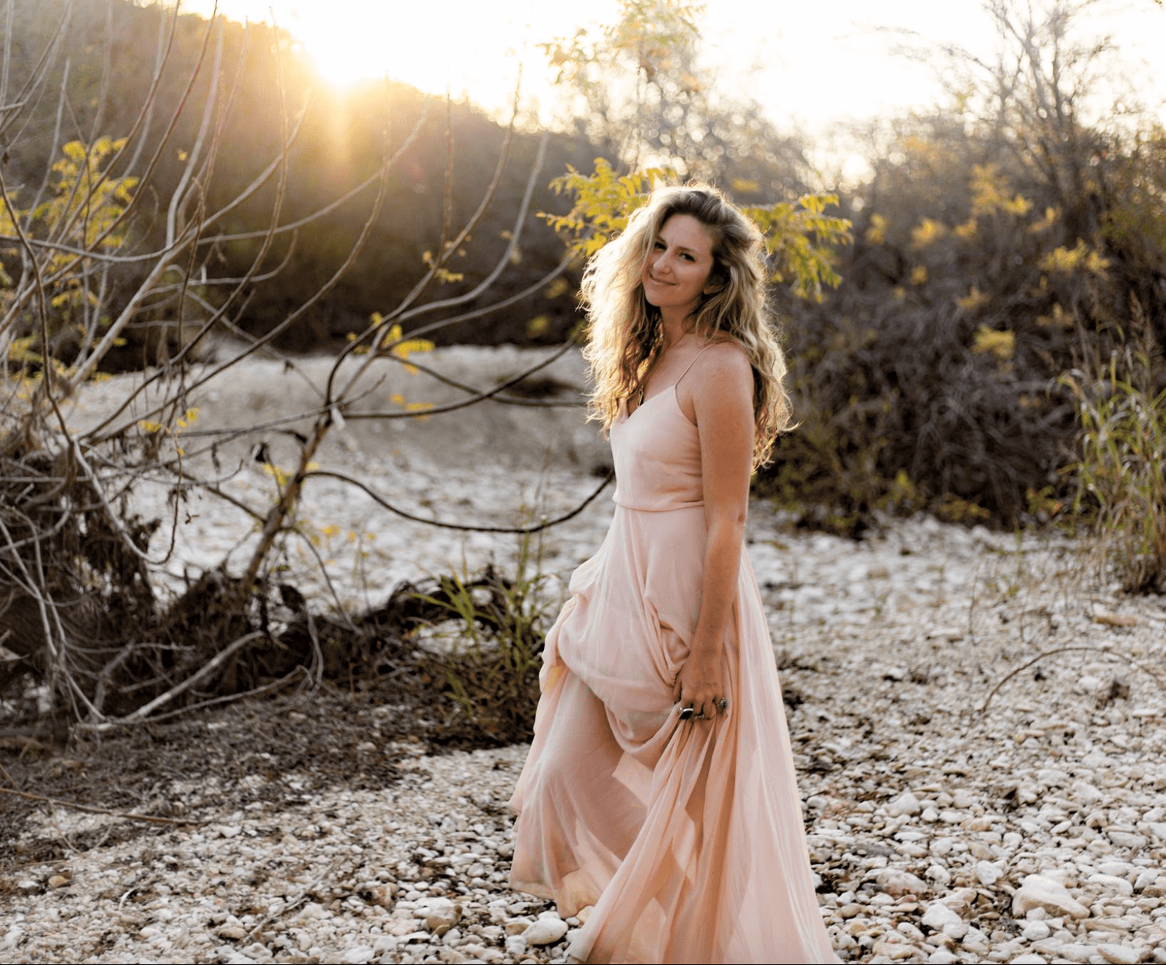 Woman in a flowing pink dress standing outdoors near a river with trees and sunlight in the background.