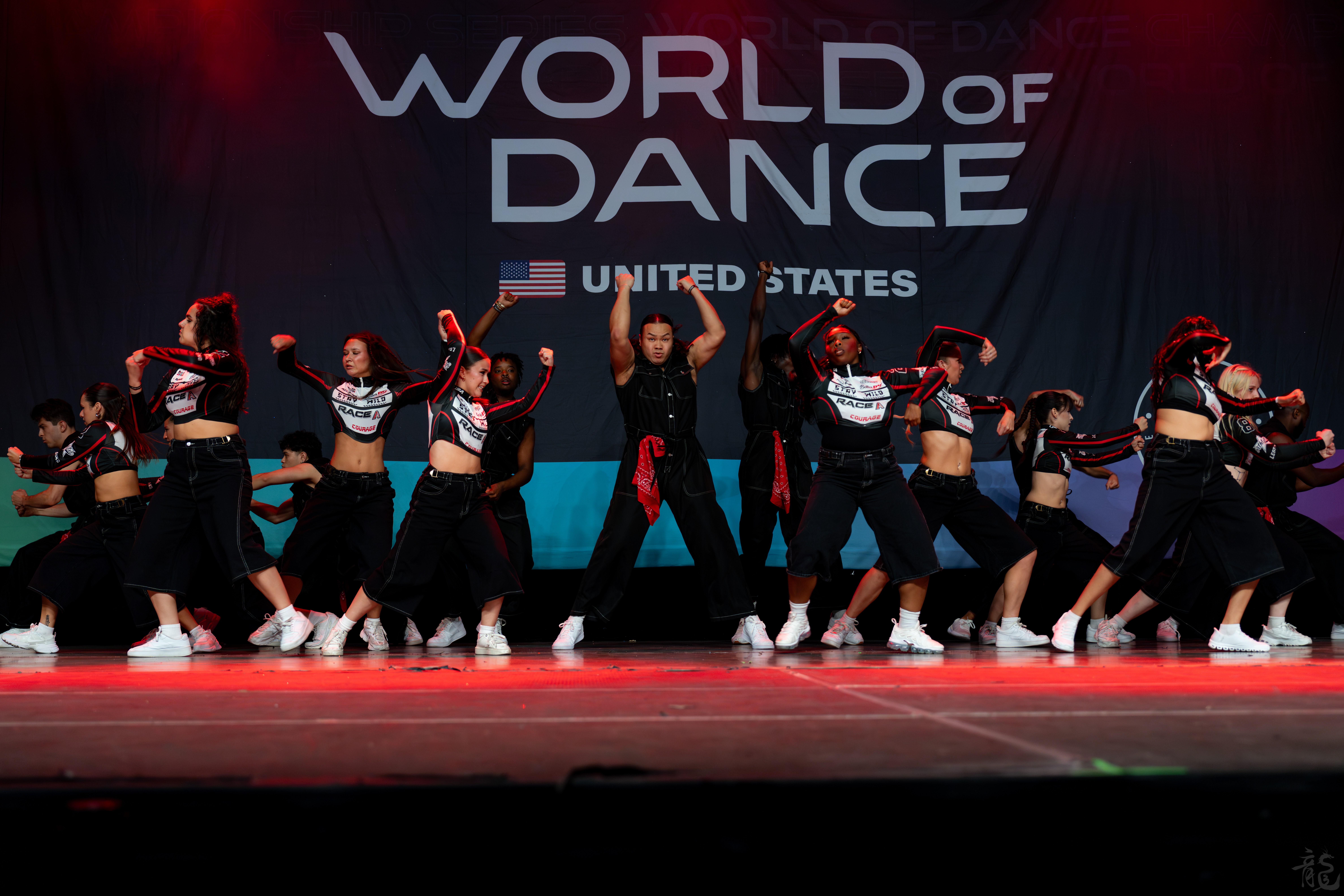 Group of young dancers performing on stage with a large 'World of Dance' banner in the background, stage lit with red and blue lights.