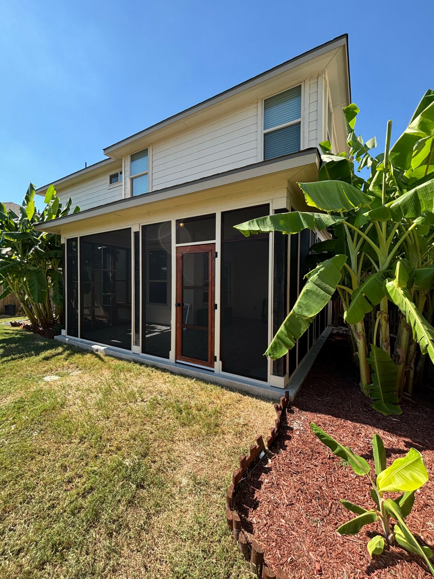 Two-story house with screened porch, surrounded by green plants and a lawn, under a clear blue sky.