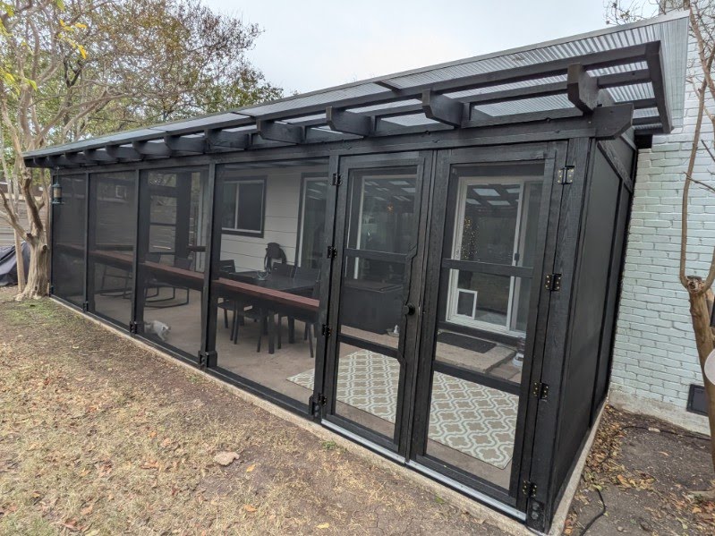 Long black enclosed porch with glass doors and windows, outdoor setting with trees and ground, metal roof, and interior visible.