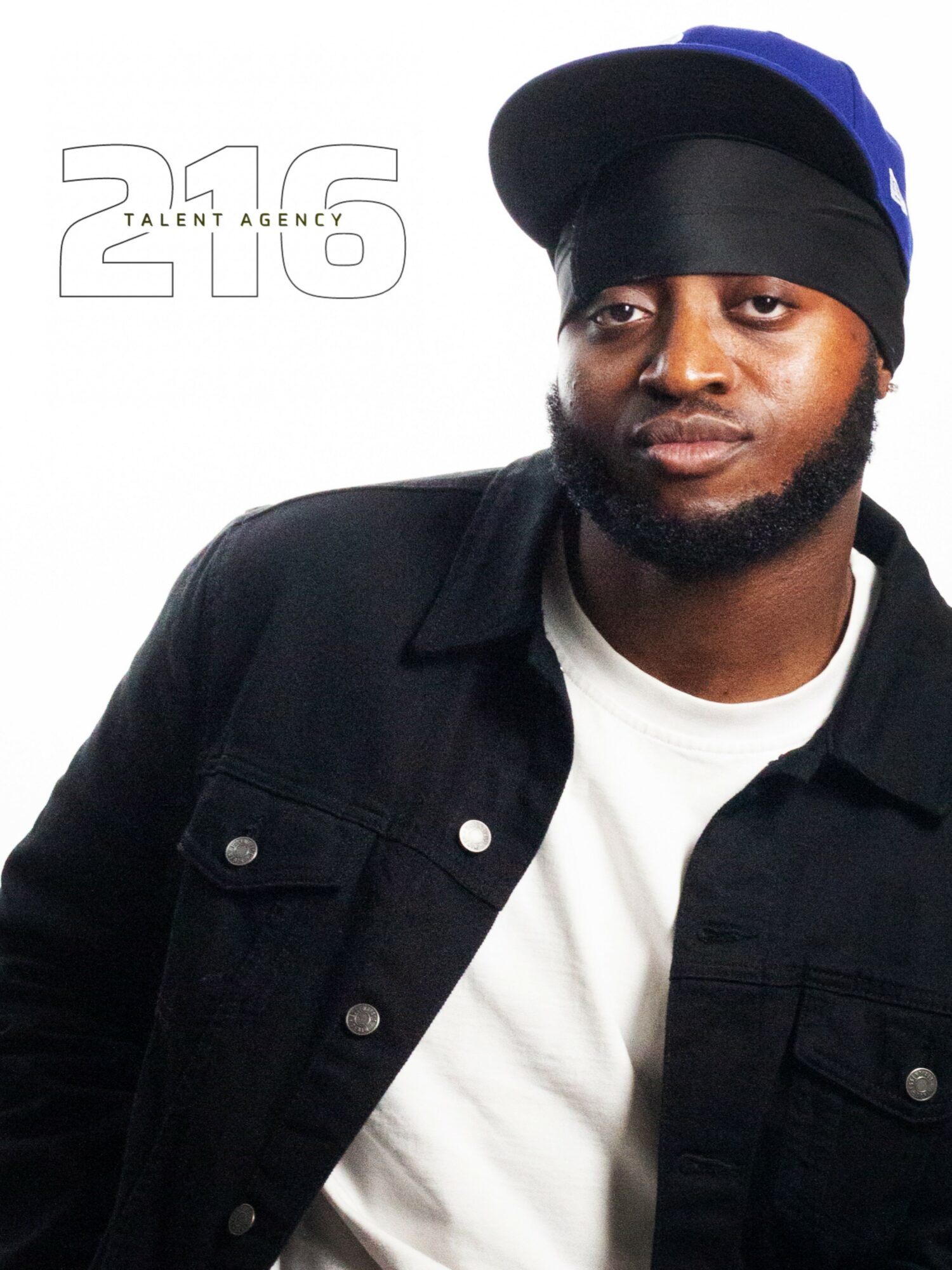 Portrait of a man wearing a black jacket, white shirt, and a black cap with a blue logo, looking at the camera.