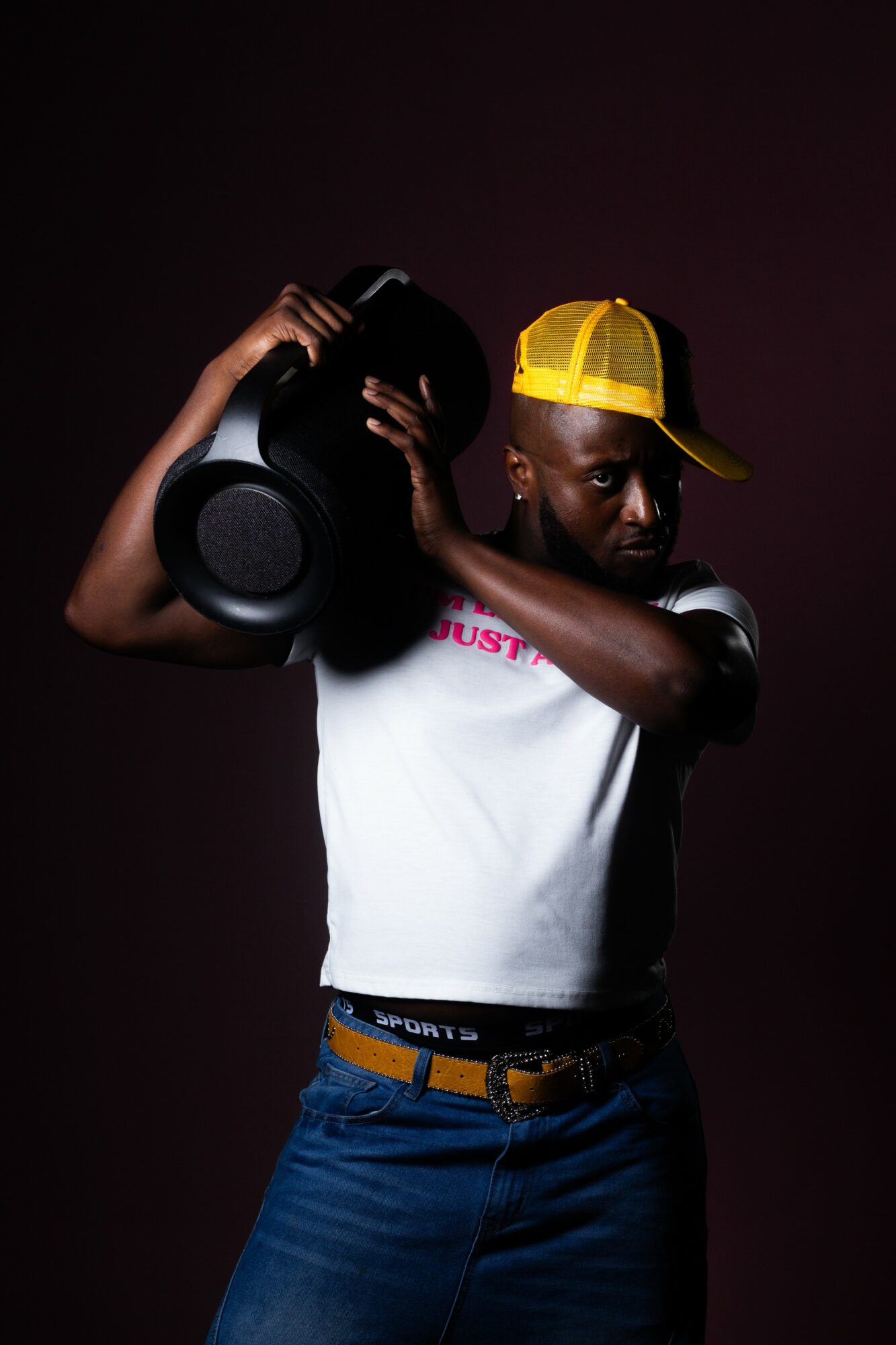 Person wearing a yellow cap, white shirt, and blue jeans holding a boombox on shoulder against dark background.