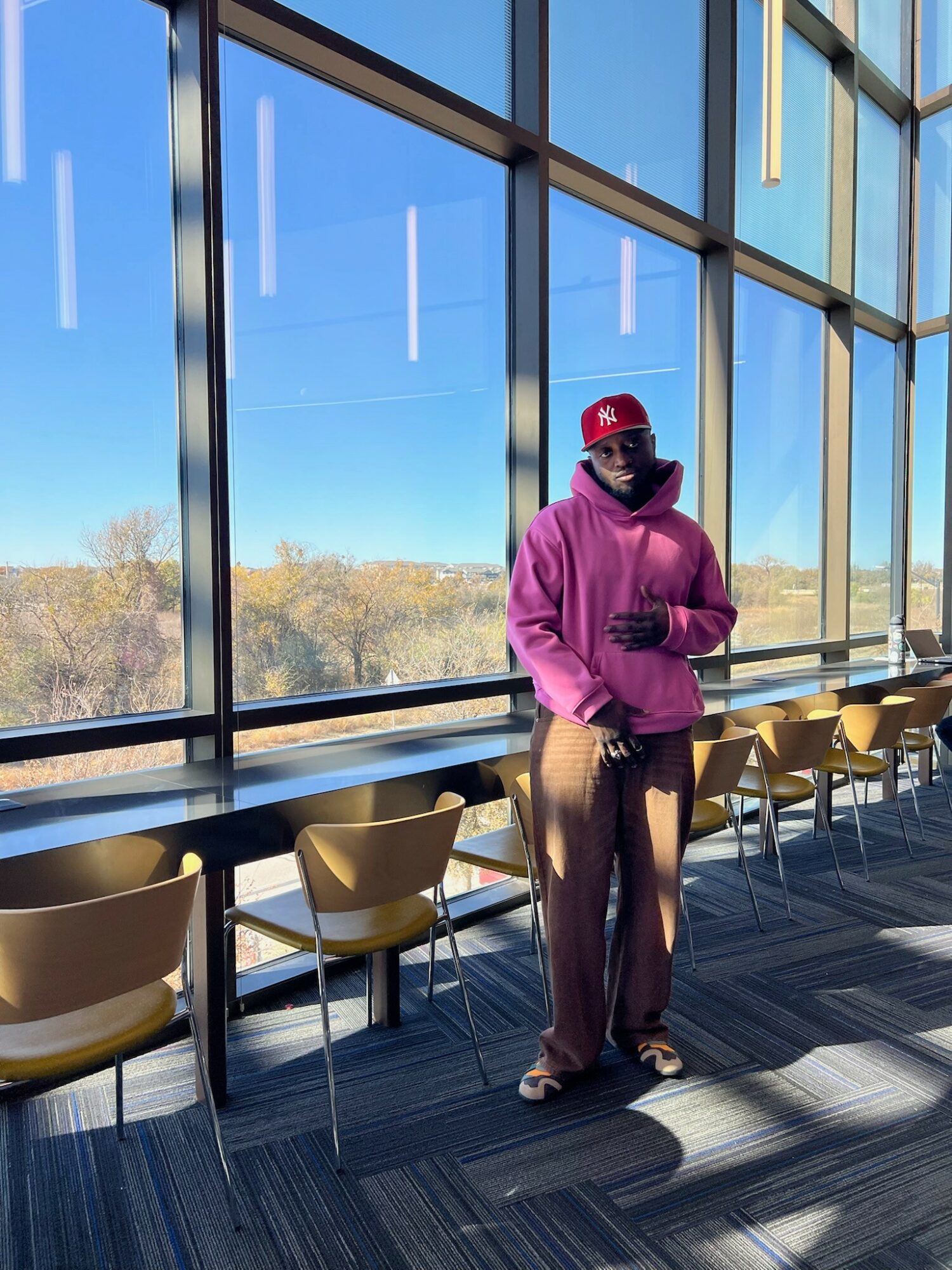 Person in purple hoodie, red cap, and patterned pants standing near large glass windows with chairs and a view outside.