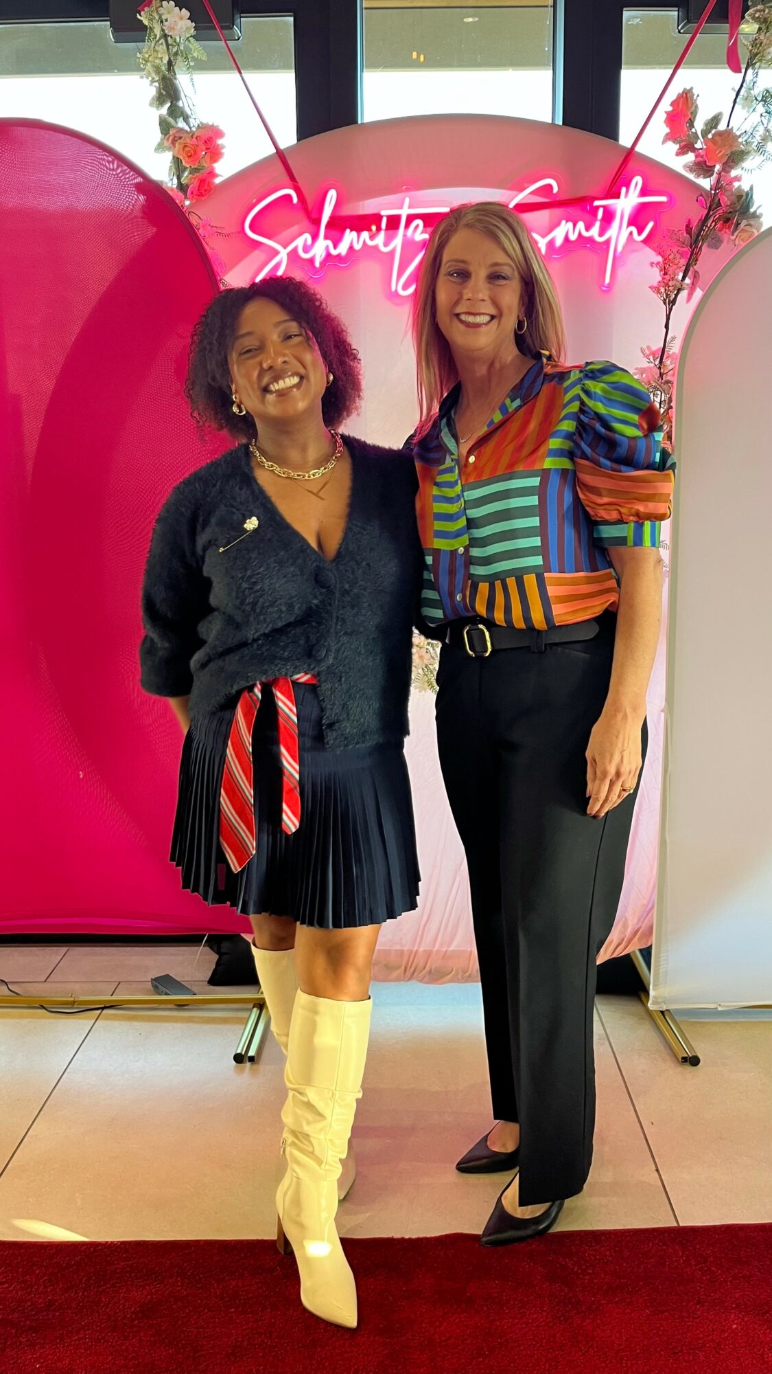 Two women stand together in front of a pink backdrop with neon sign, smiling, one wearing a denim jacket and yellow boots.